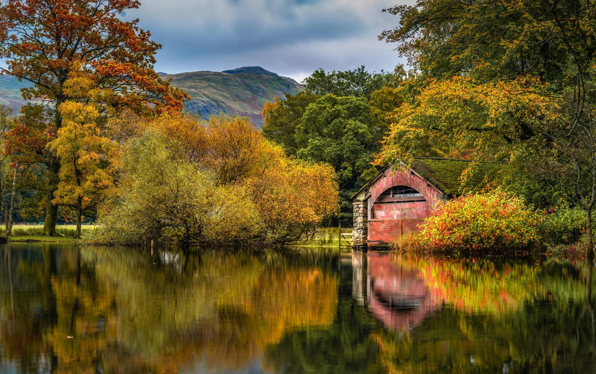 Scenic autumn landscape with a red boathouse beside a lake, surrounded by colorful trees reflecting in the water.