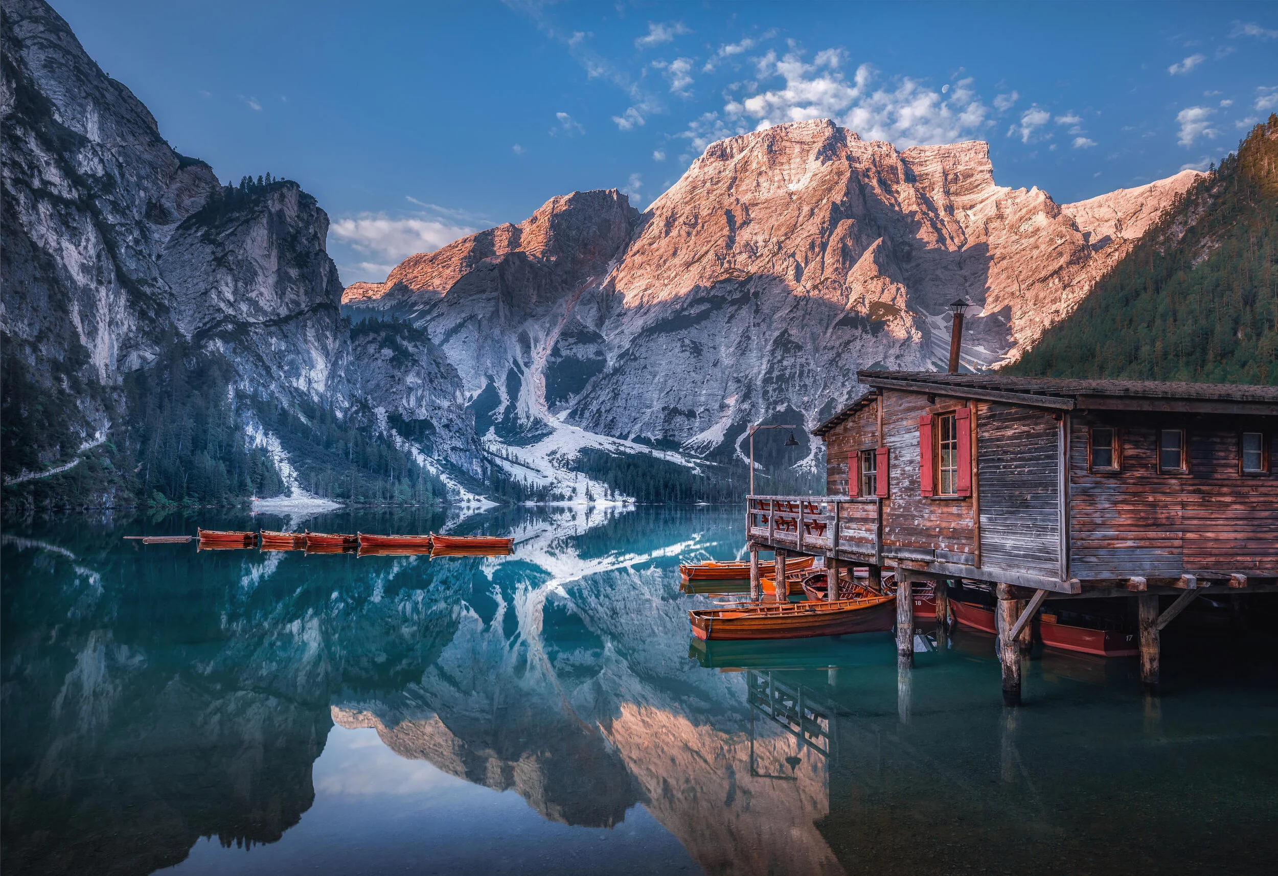 A serene mountain lake reflecting surrounding peaks, a wooden boathouse, and moored boats.