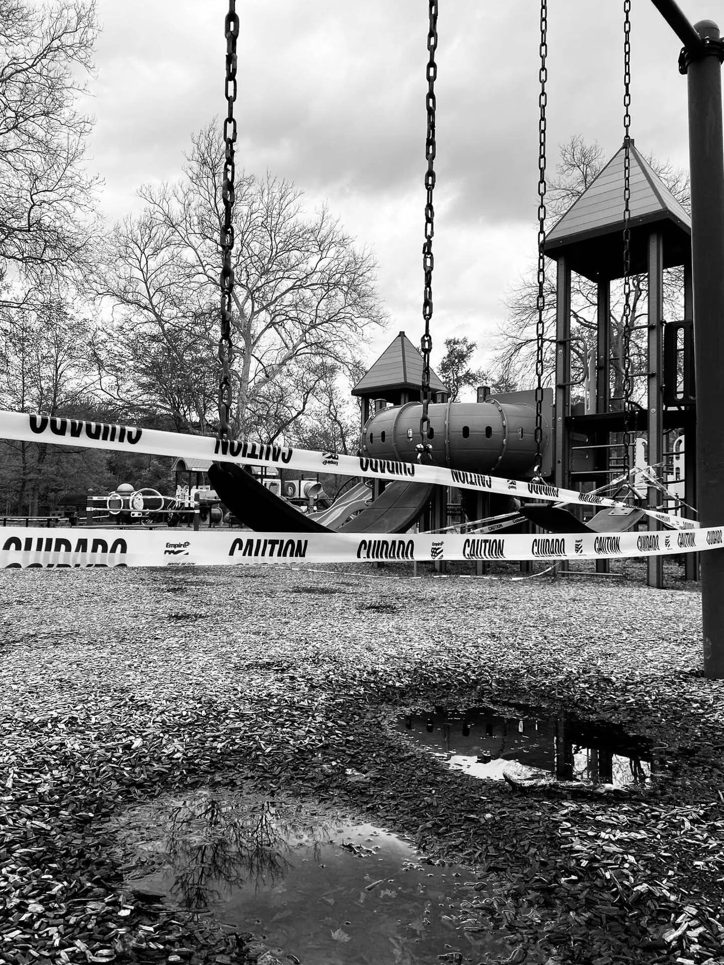 Black and white photo of a playground with swings and play structures, surrounded by caution tape. The ground is wet with visible puddles, and trees without leaves are in the background.