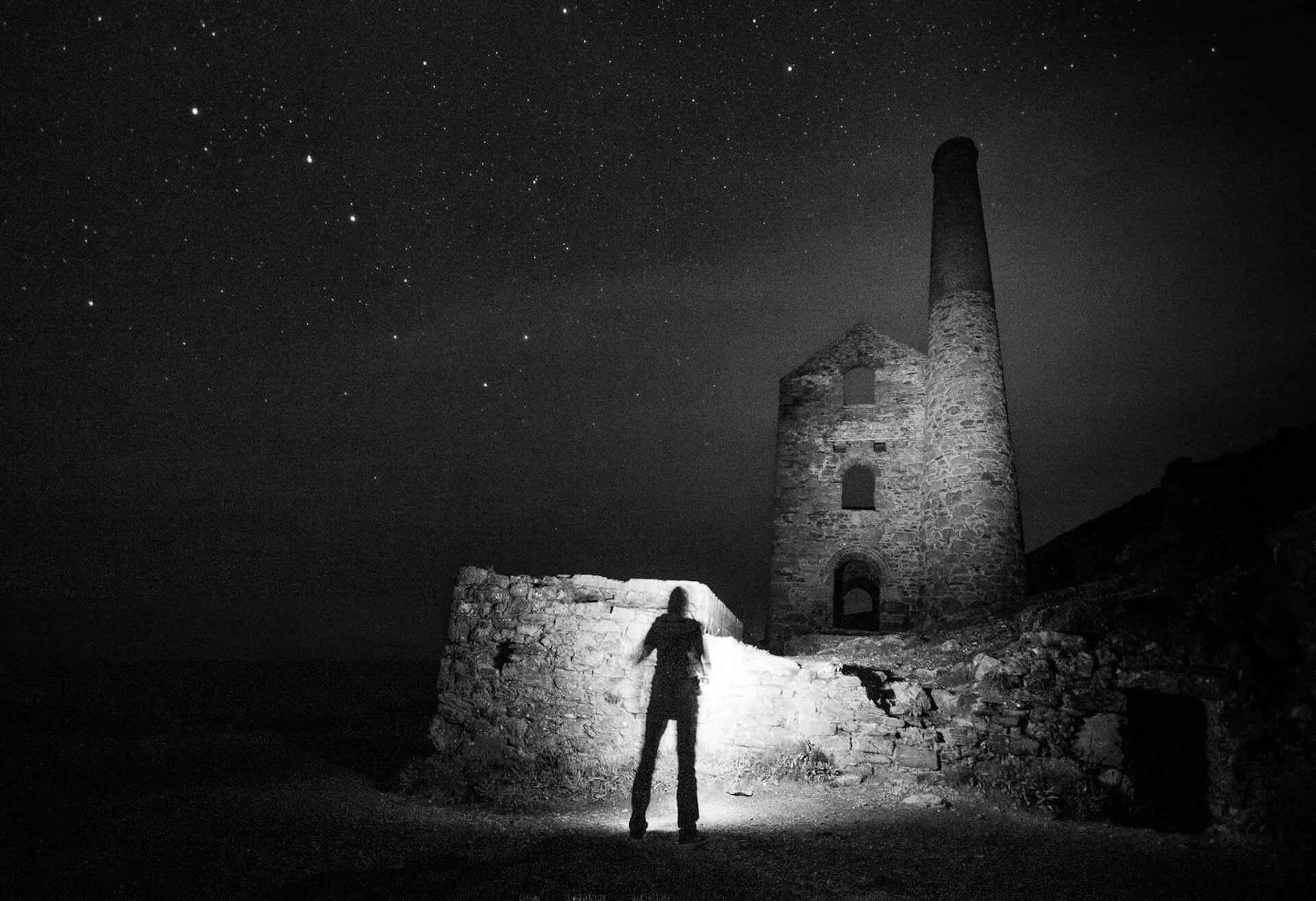 Silhouette of a person standing in front of an old stone building with a tall chimney at night, under a starry sky. The scene is illuminated by a flashlight or torch.