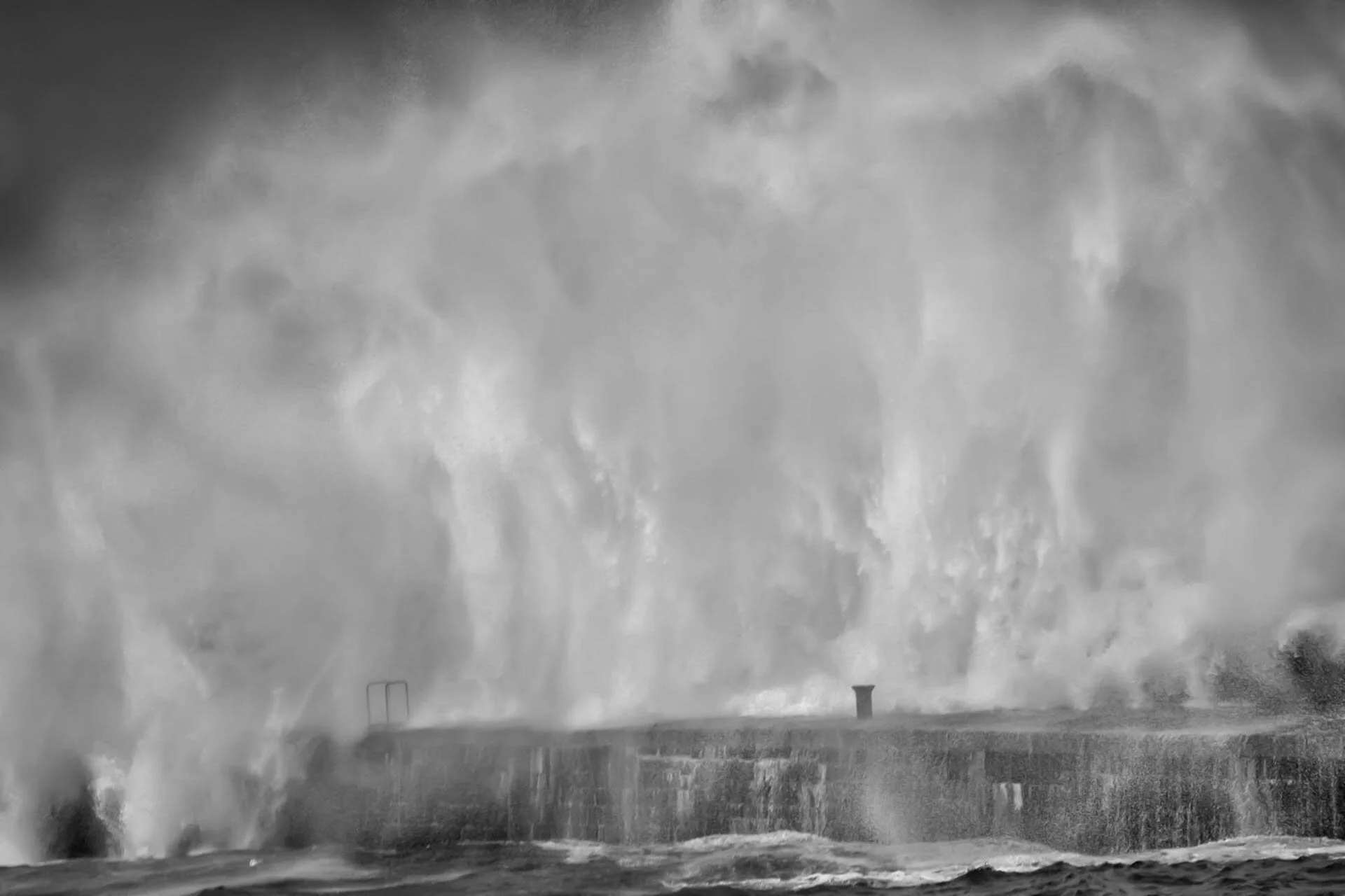 Huge wave crashing against a sea wall, creating a spray in black and white.