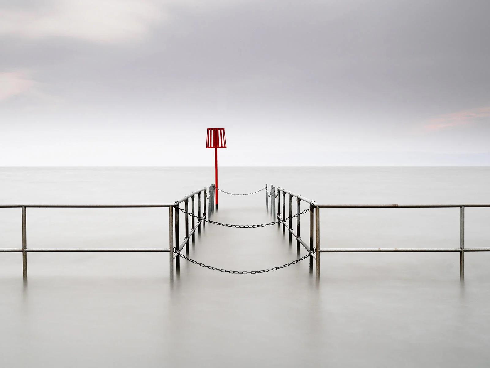 A minimalist seascape featuring a metal railing with chains extending into calm, blurred water, under a gray sky. A red navigational post stands at the end of the railing structure.