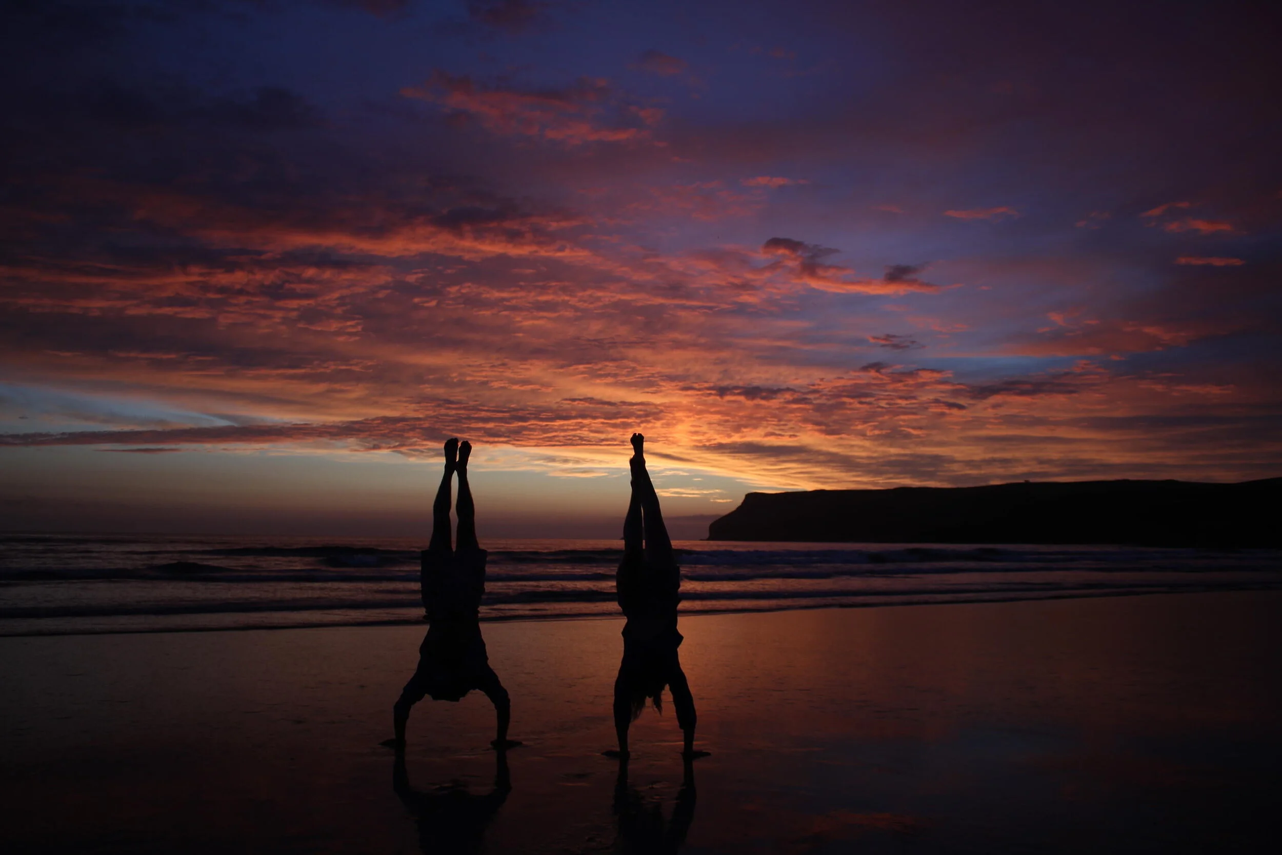 Two people doing handstands on a beach during a colorful sunset.