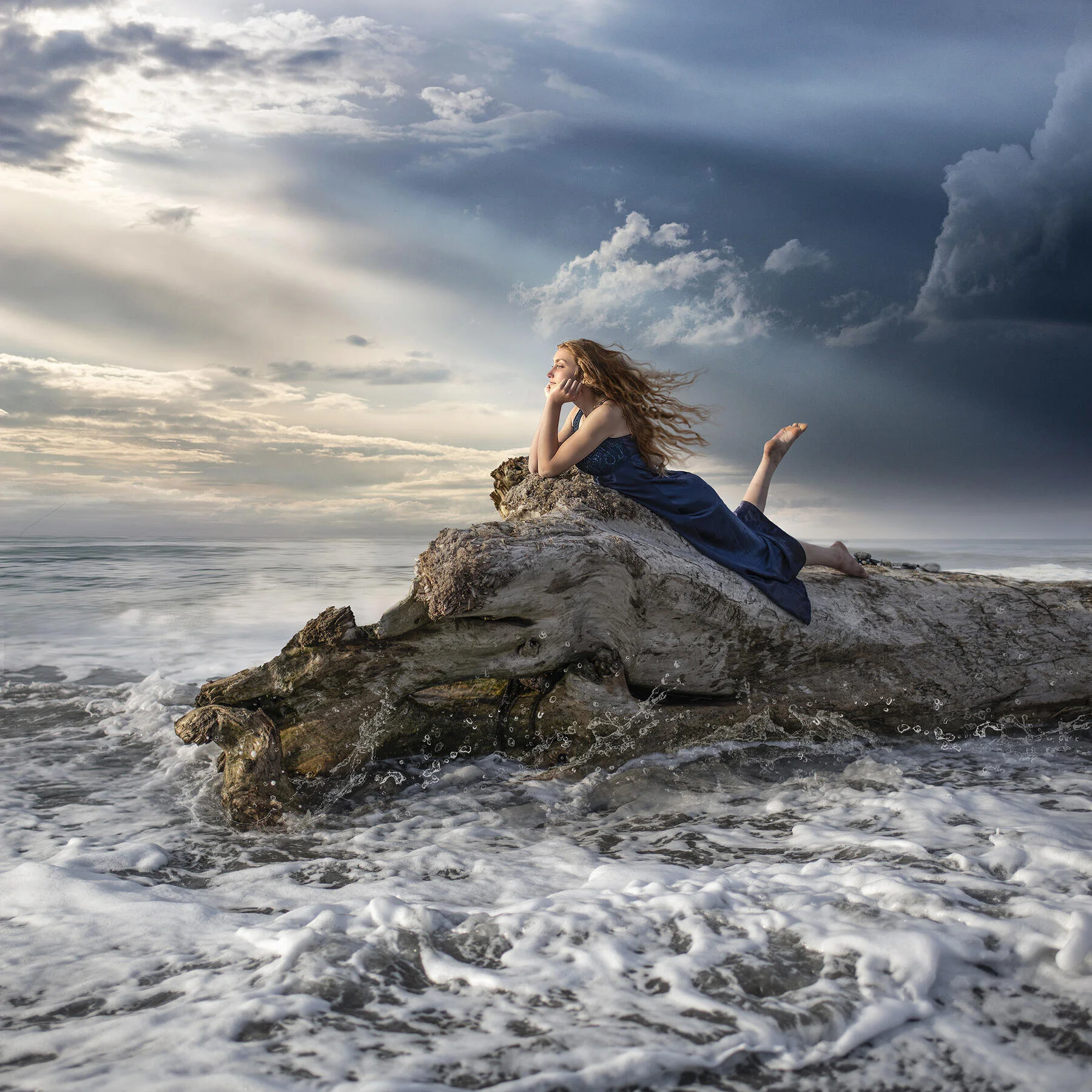A woman in a blue dress is lying on a large driftwood log by the ocean, with waves crashing around her and a dramatic cloudy sky in the background.