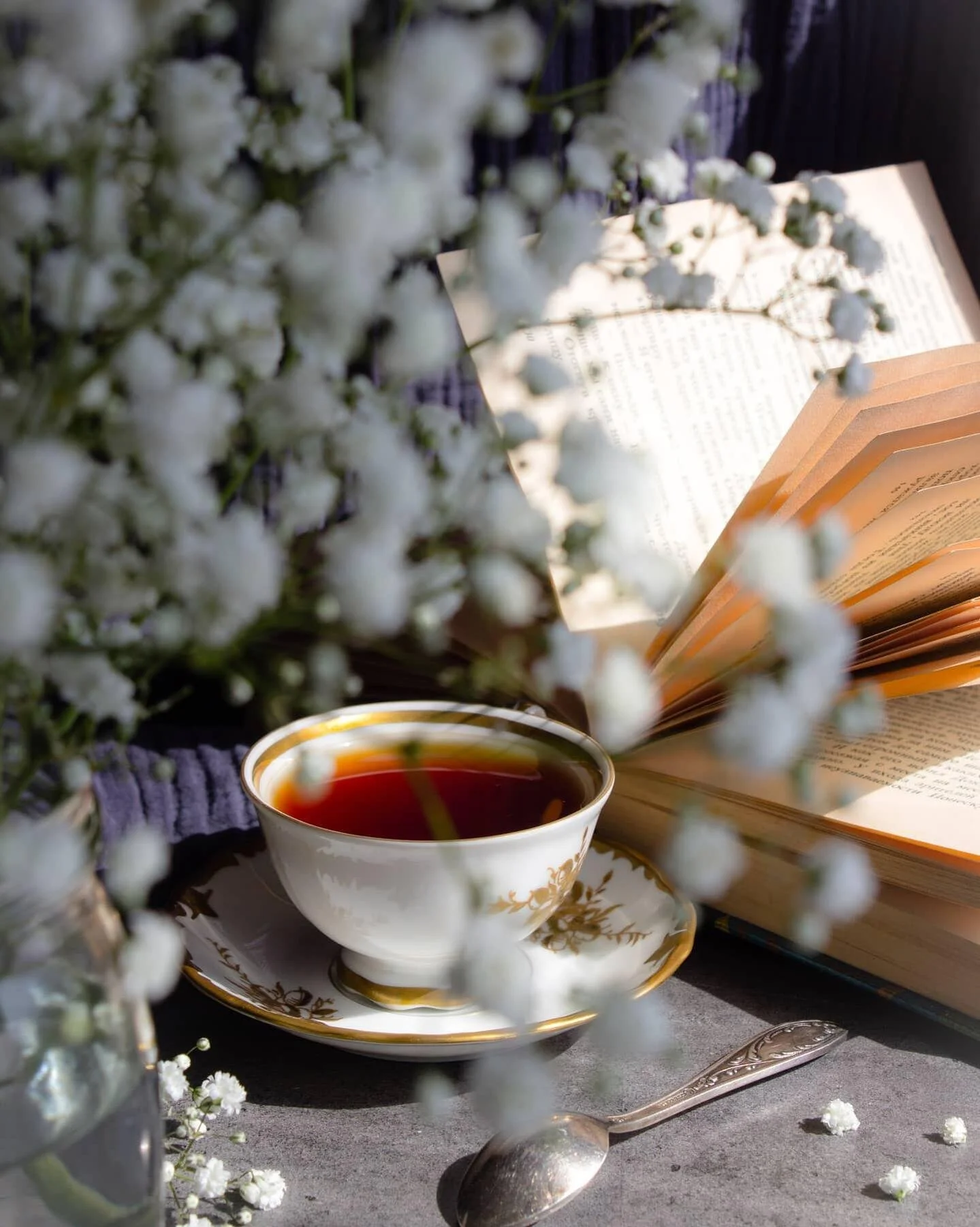 Cup of tea on a saucer in front of an open book, surrounded by small white flowers and a spoon, with a soft-focus effect.