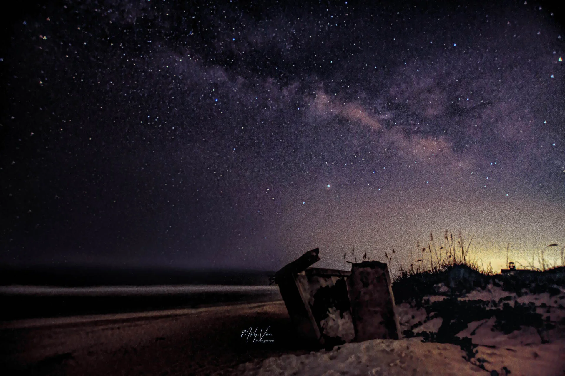 Starry night sky over a beach with sand dunes and sparse vegetation in the foreground.