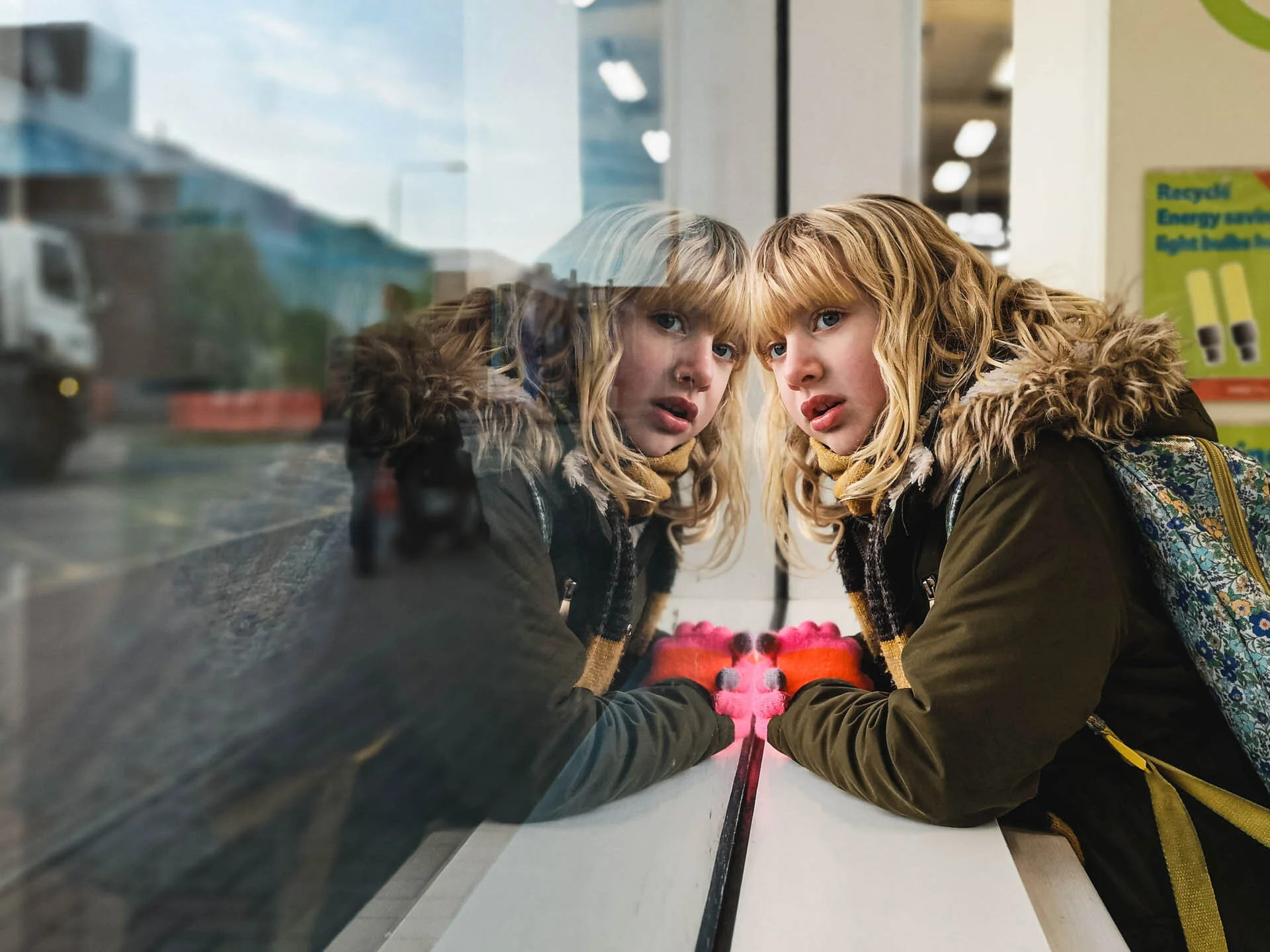 Young person with blonde hair looking into a window with reflection, wearing pink gloves and a green coat with fur collar, city street view outside.