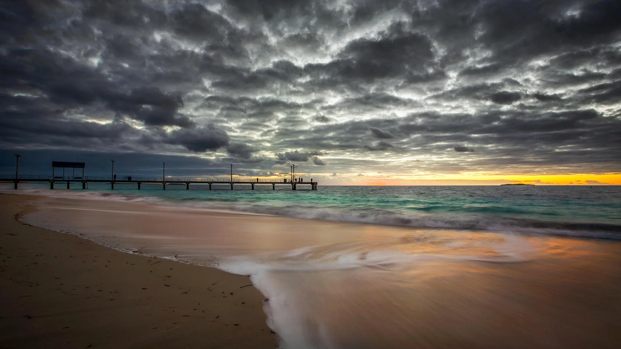 Beach scene with a pier and cloudy sky during sunset, featuring waves on the shore.