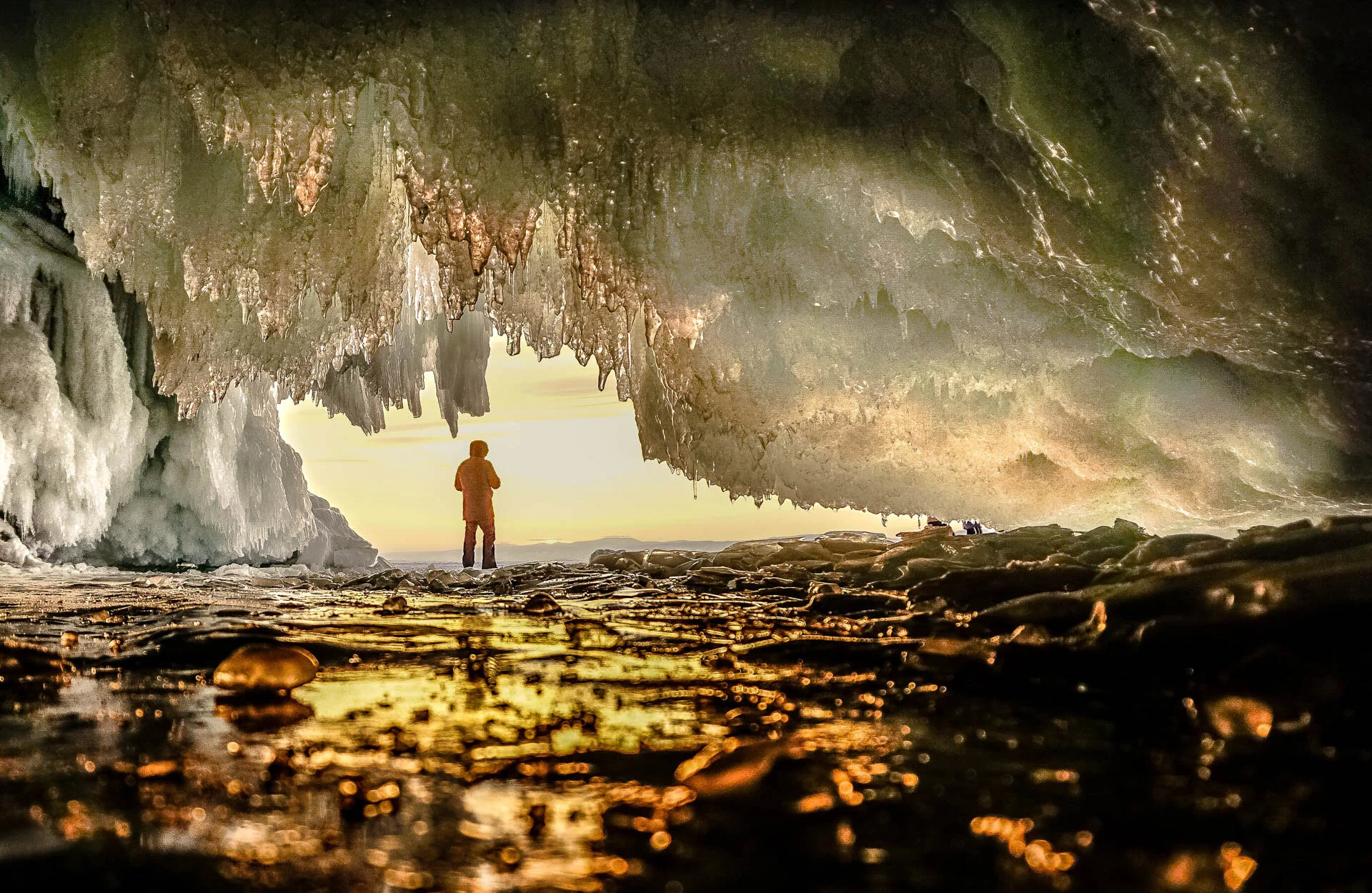 Person standing inside an ice cave with icicles, looking out at a sunset.