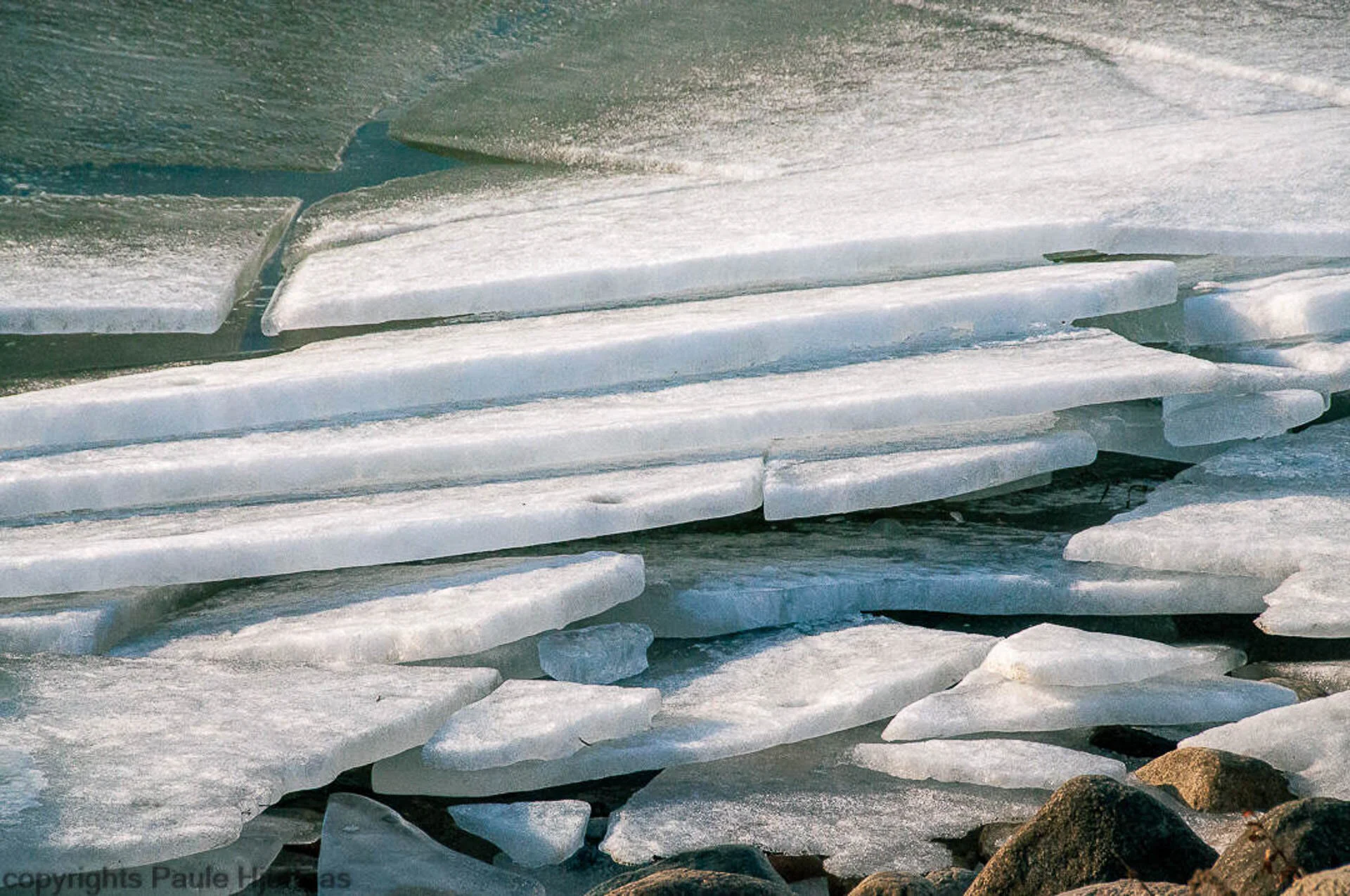 Stacked ice sheets on a frozen lake with rocks at the edge.