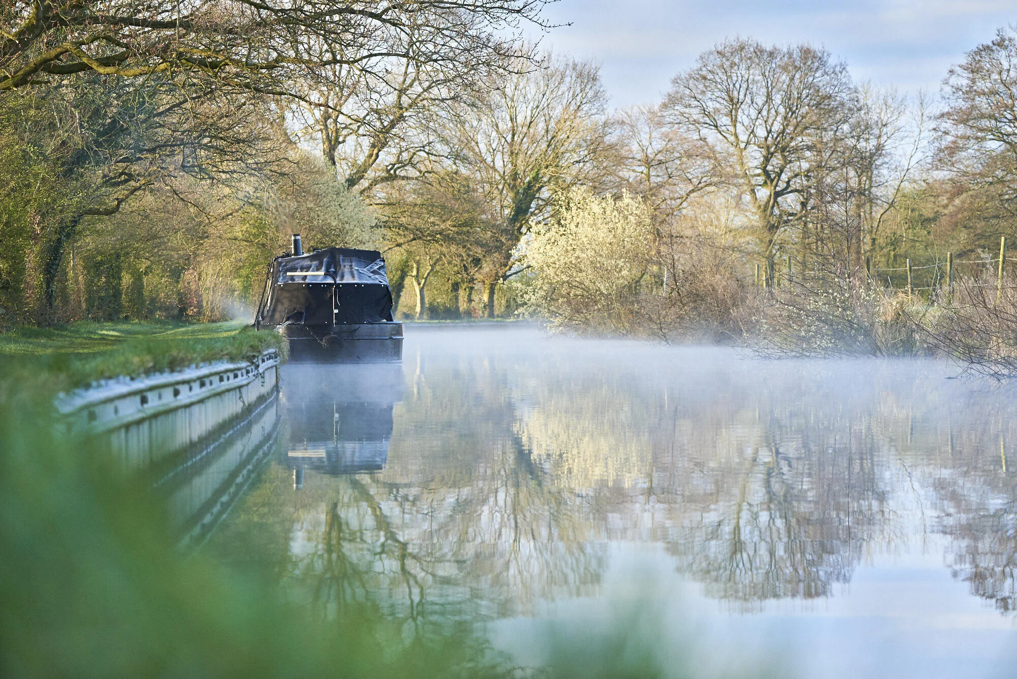 Narrowboat on a calm canal surrounded by leafless and blossoming trees, with mist rising from the water.