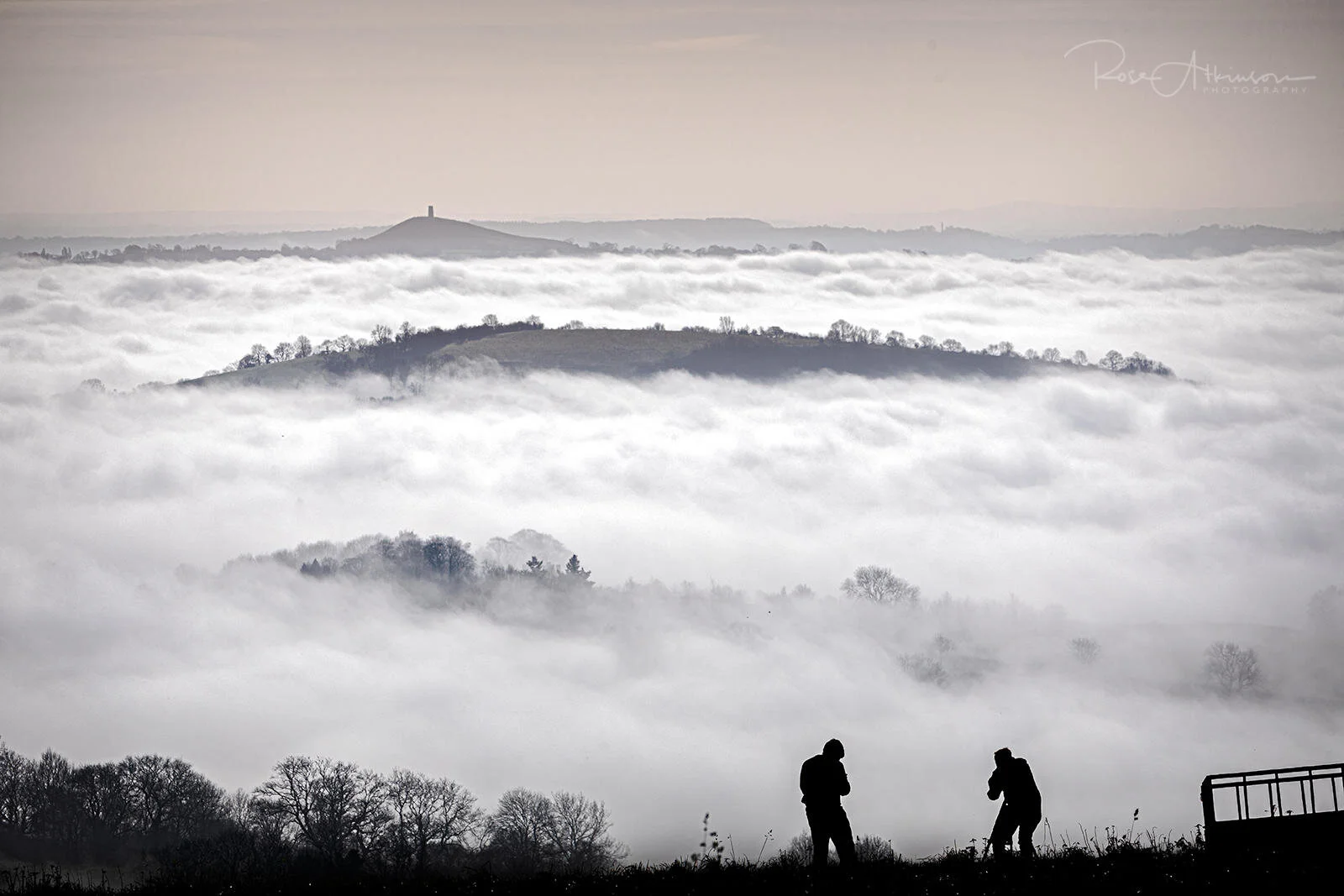 Silhouettes of two people on a hill with fog-covered landscape and distant mound visible