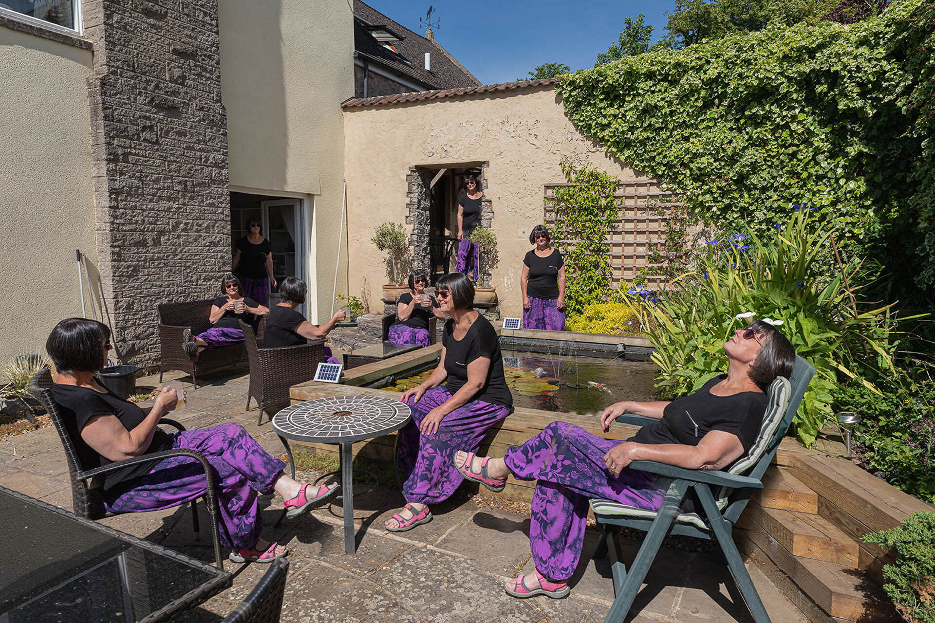 A garden scene with multiple duplicated images of a woman wearing a black shirt and purple pants, sitting and standing in various positions. The setting includes patio furniture, a pond, and greenery.
