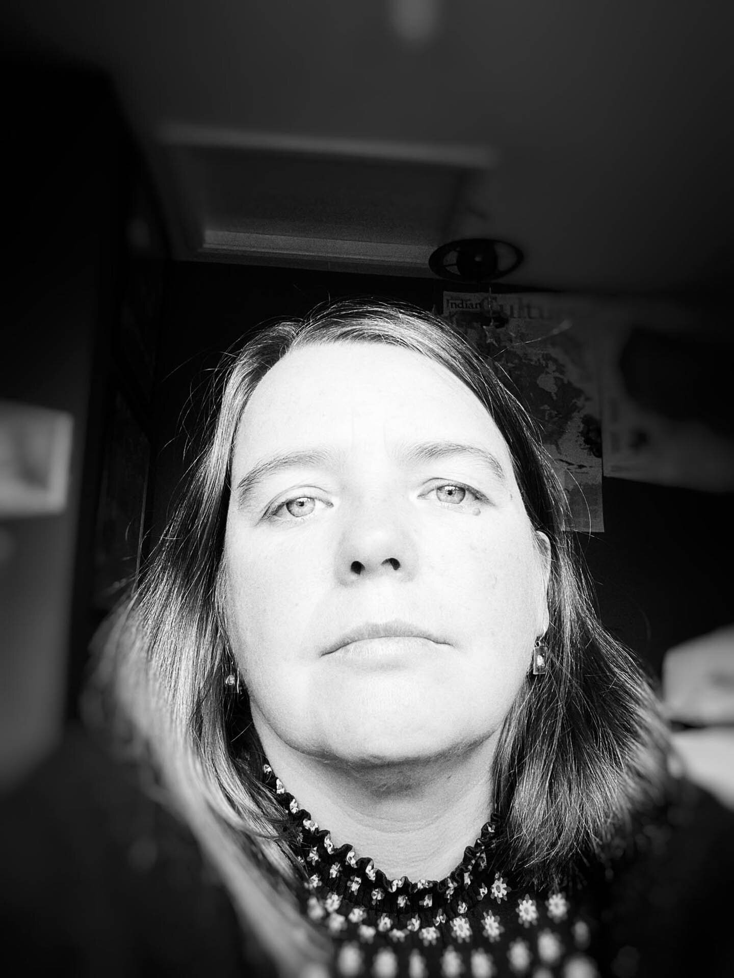 Black and white portrait of a woman with straight hair, wearing a patterned top, looking directly at the camera, with a blurred background.