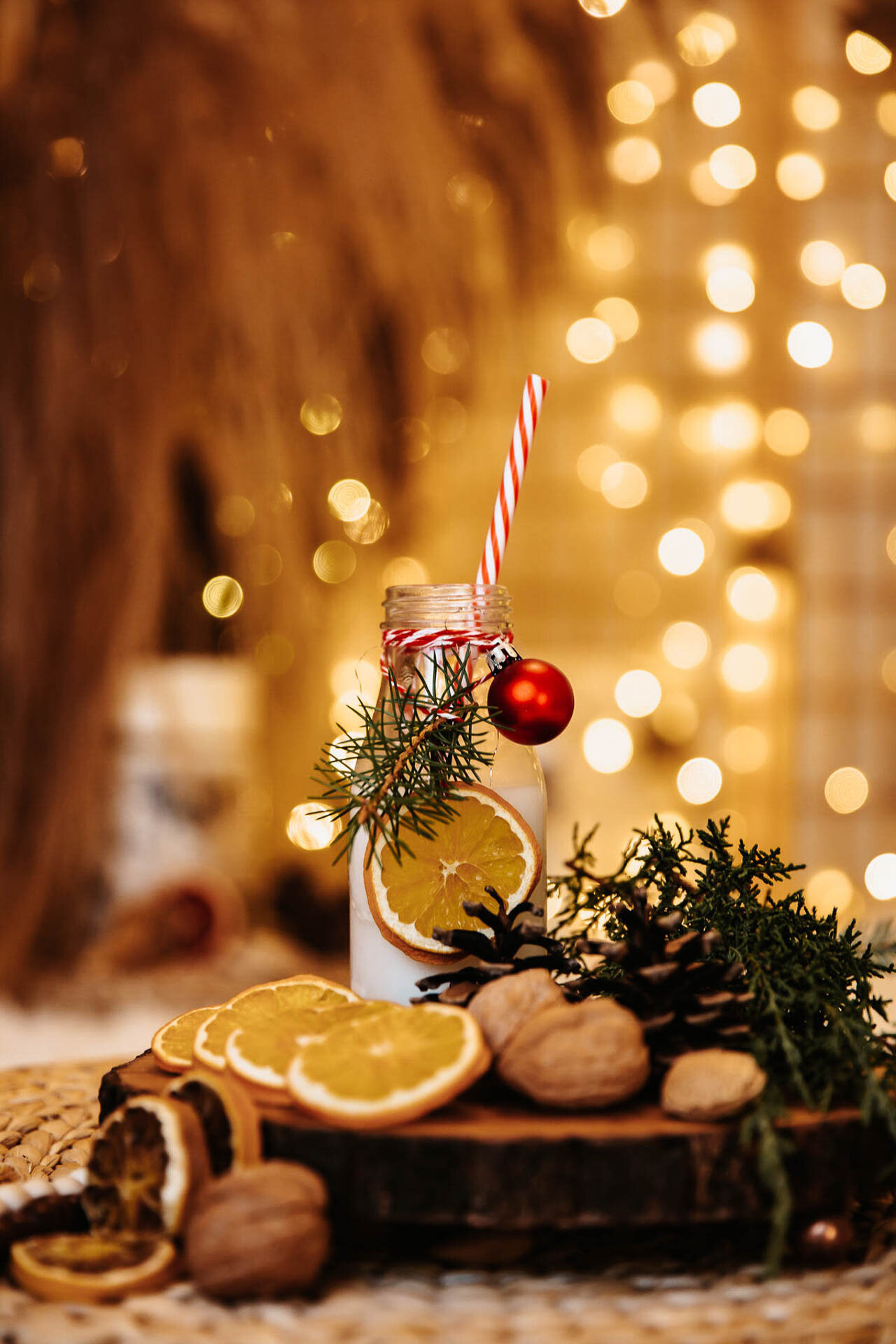 Festive arrangement with milk bottle, red and white straw, orange slices, pine branch, red ornament, nuts, and pinecones, against blurred lights background.