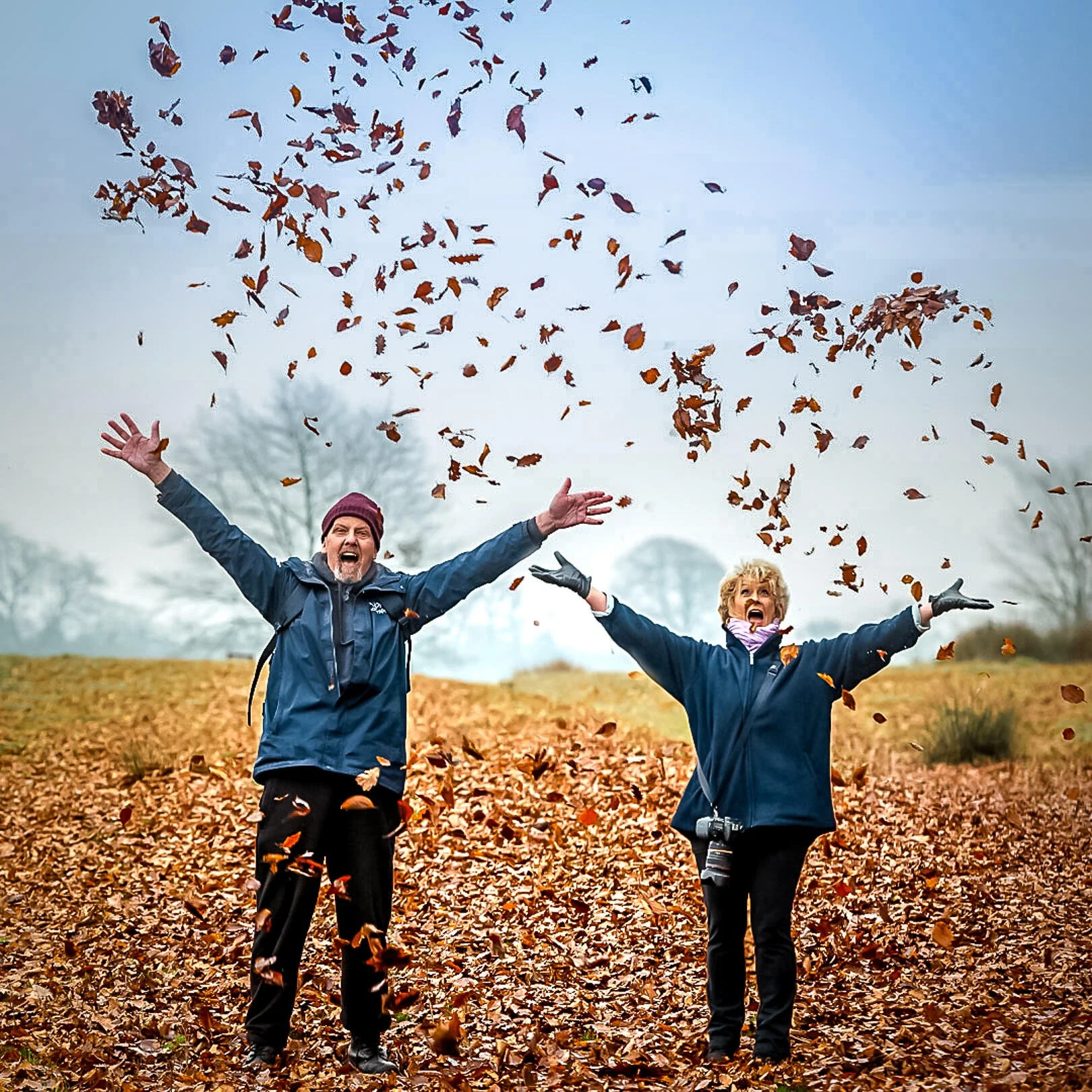 Two people joyfully throwing autumn leaves in the air in a park, wearing winter jackets and gloves.