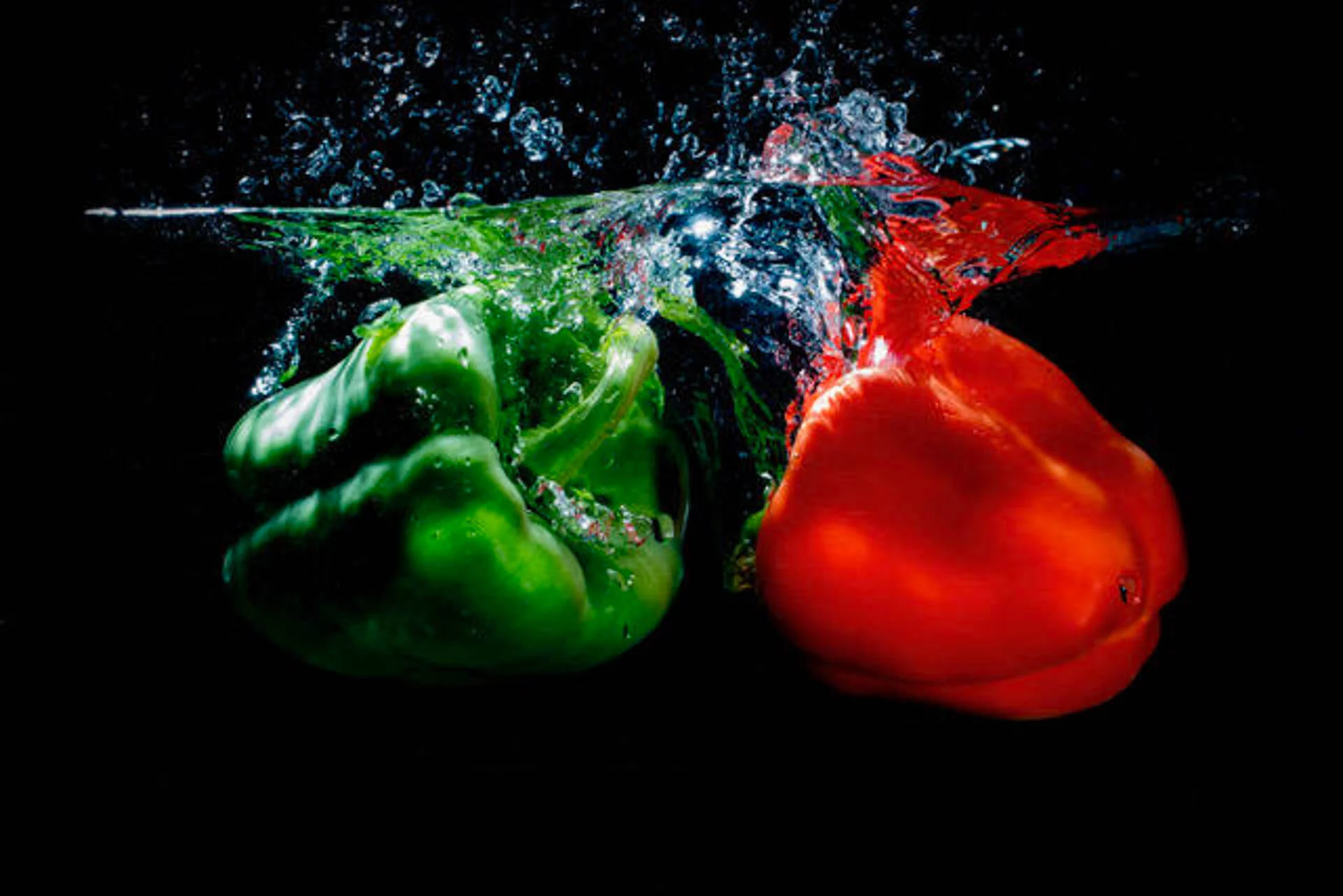 Green and red bell peppers splashing into water against a black background.
