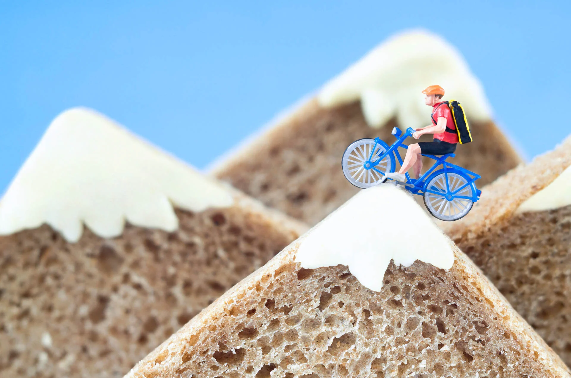 Miniature figure on a blue bicycle atop a slice of bread with cheese, mimicking a mountain scene.