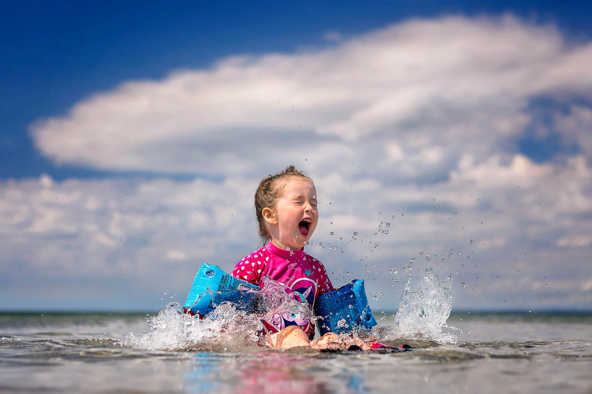 Child wearing pink swimwear and blue floaties splashing in water under a blue sky with clouds.