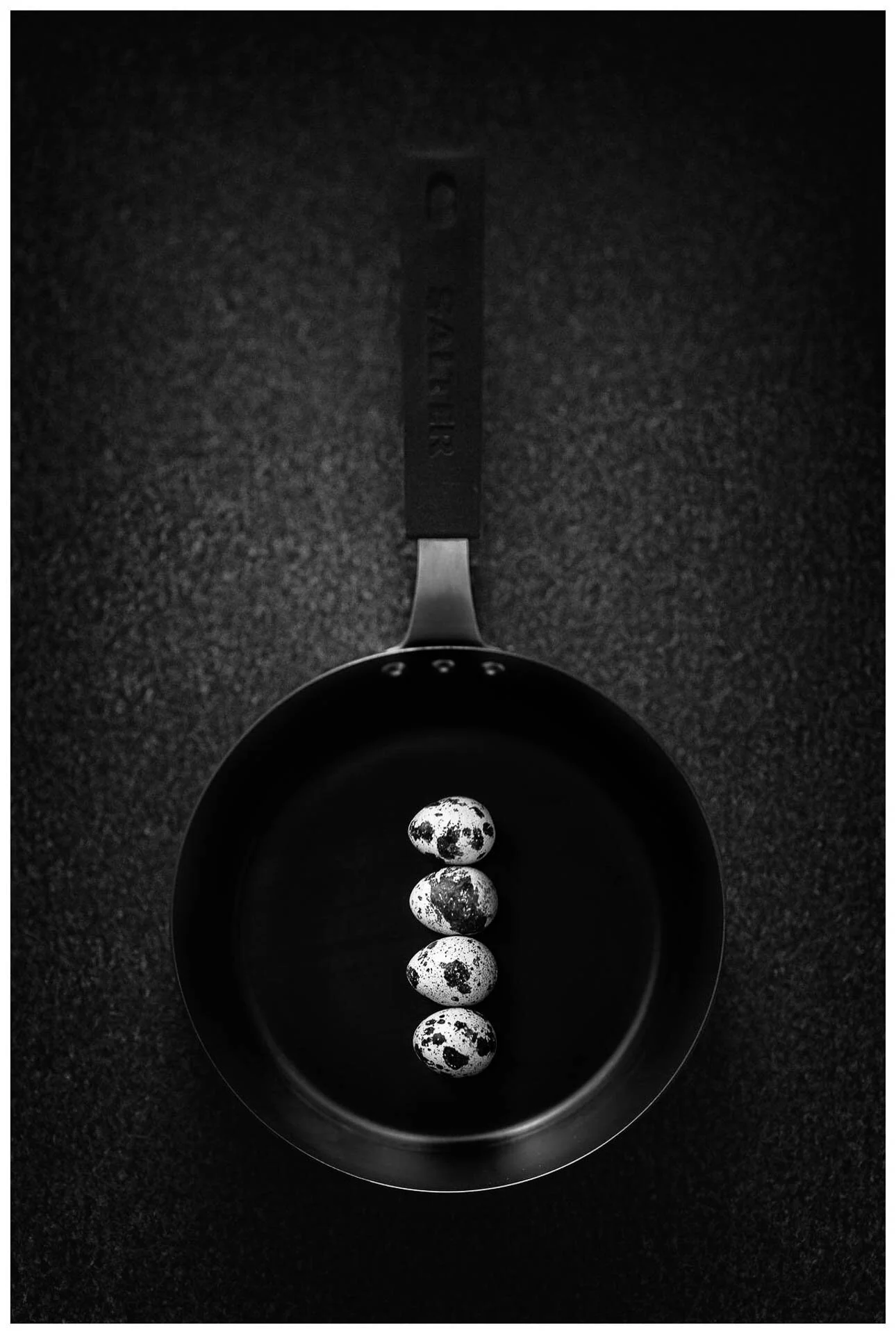 Black and white image of five quail eggs in a frying pan on a dark surface.