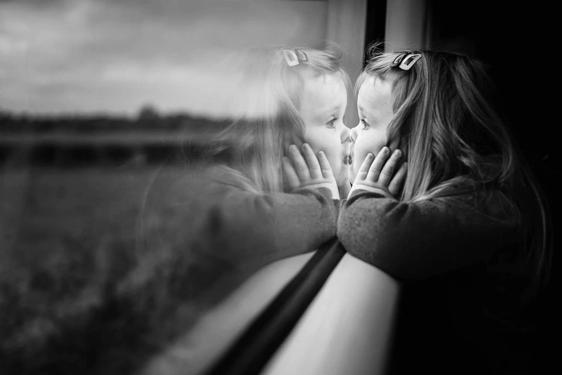 Black and white photo of a child looking out a window, with their reflection visible, hands on their cheeks, and hairclips in their hair.