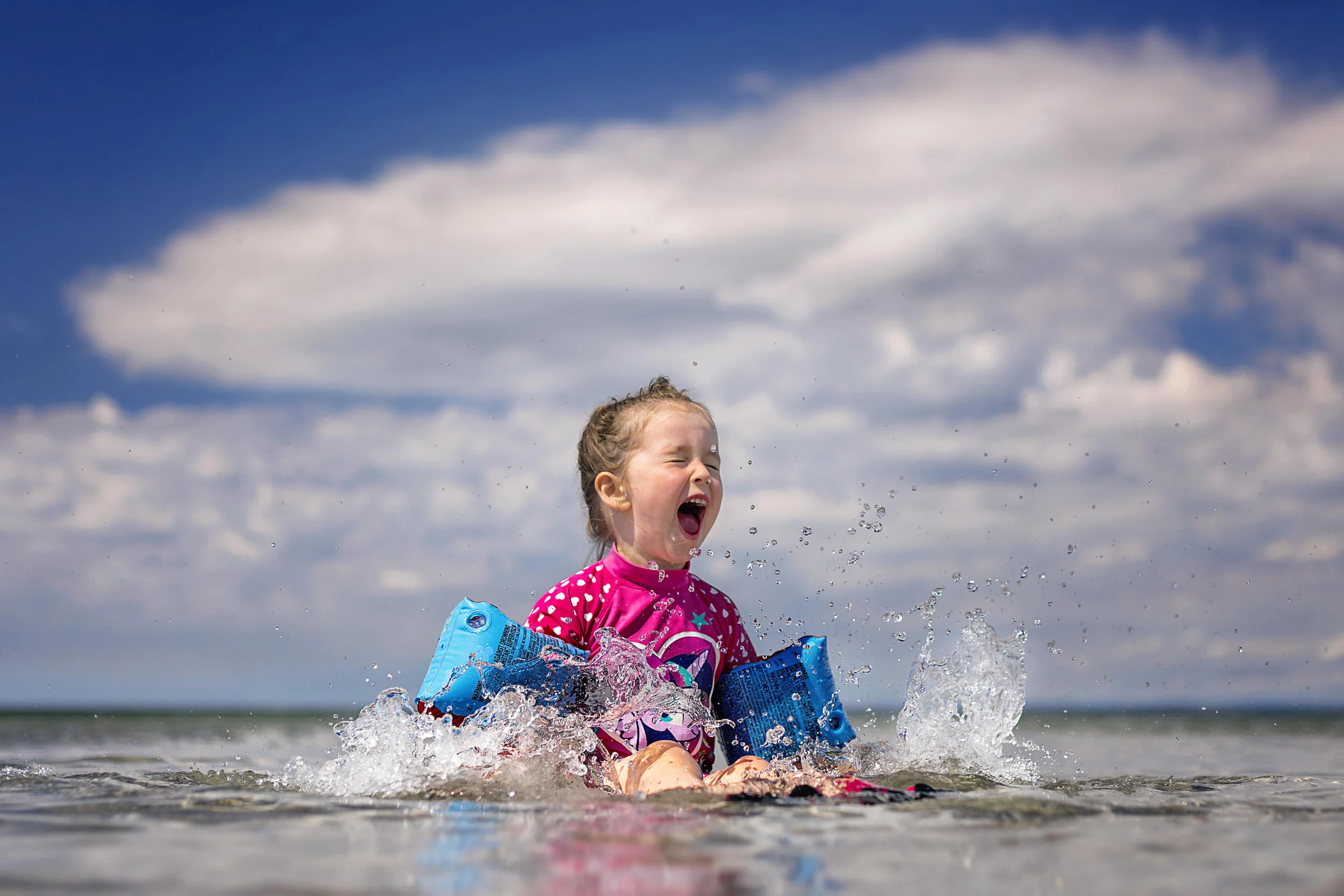 Child wearing a pink swimsuit with floaties, playing in the water, splashing, and smiling under a cloudy blue sky.