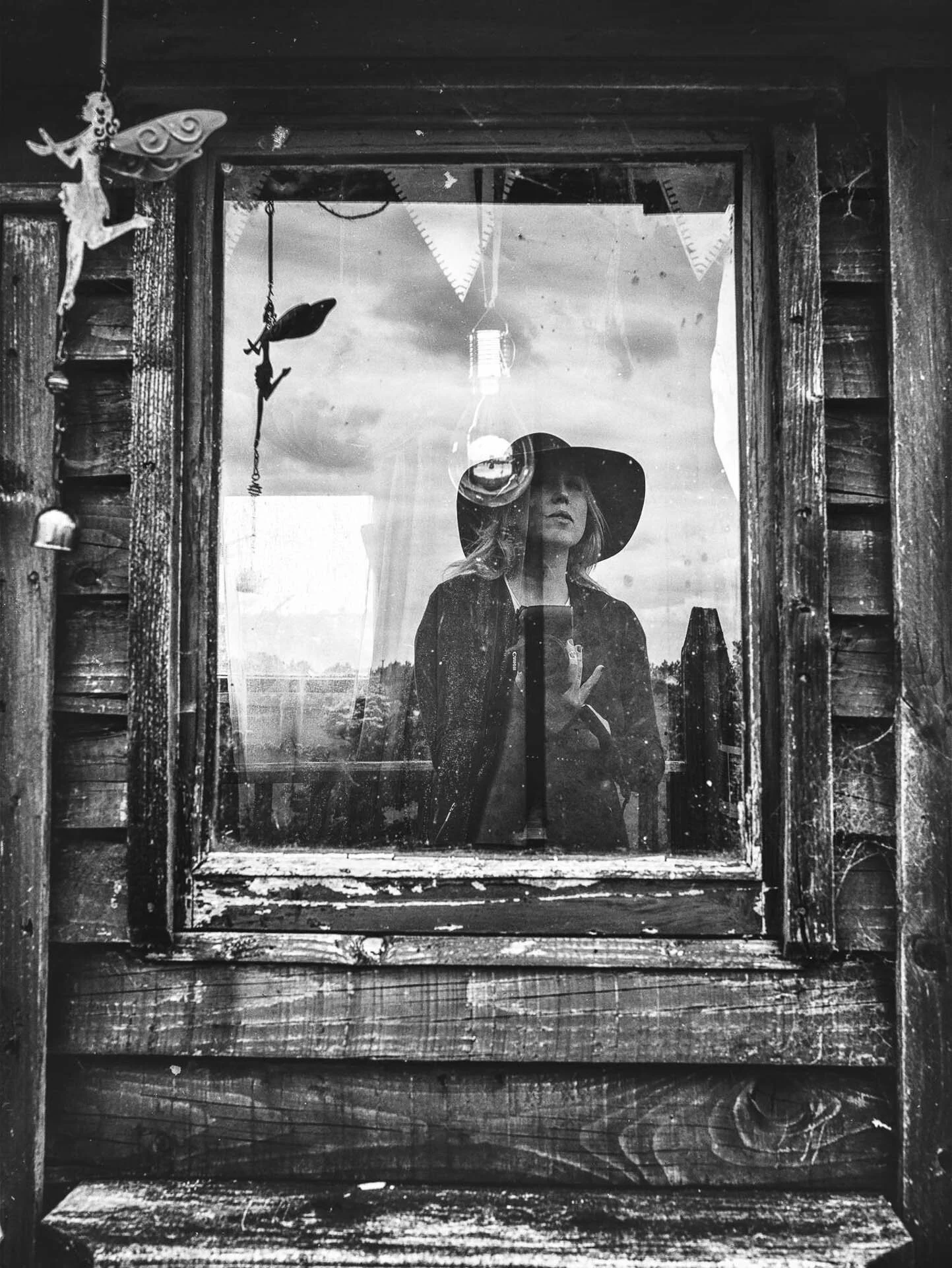 Black and white photo of a person with a hat standing behind a window, looking out. The window has a rustic wooden frame, and there are fairy decorations hanging. The reflection shows the sky and clouds.