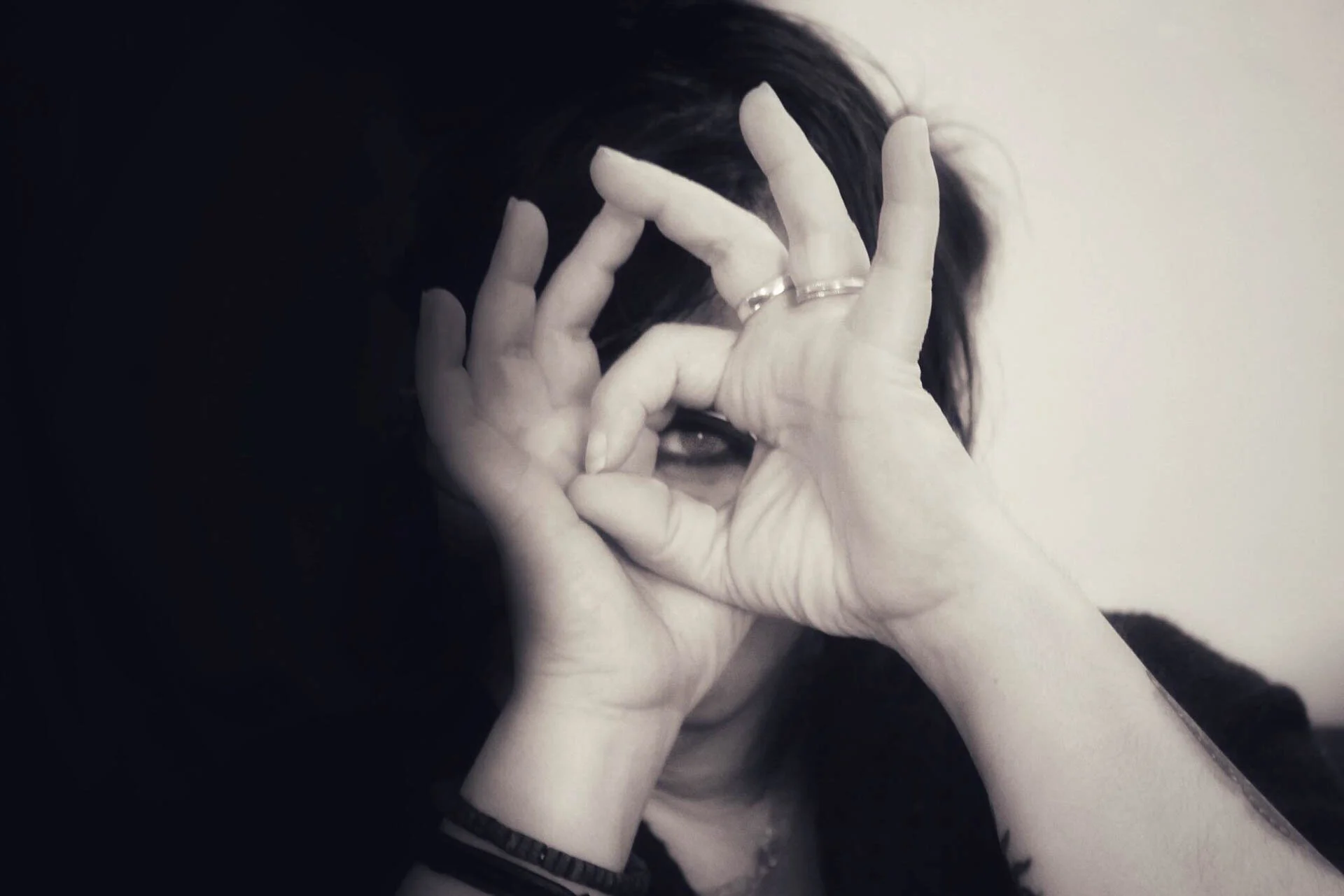 Monochrome photo of a person making a circle with their fingers, peering through it like a telescope, against a dark background.