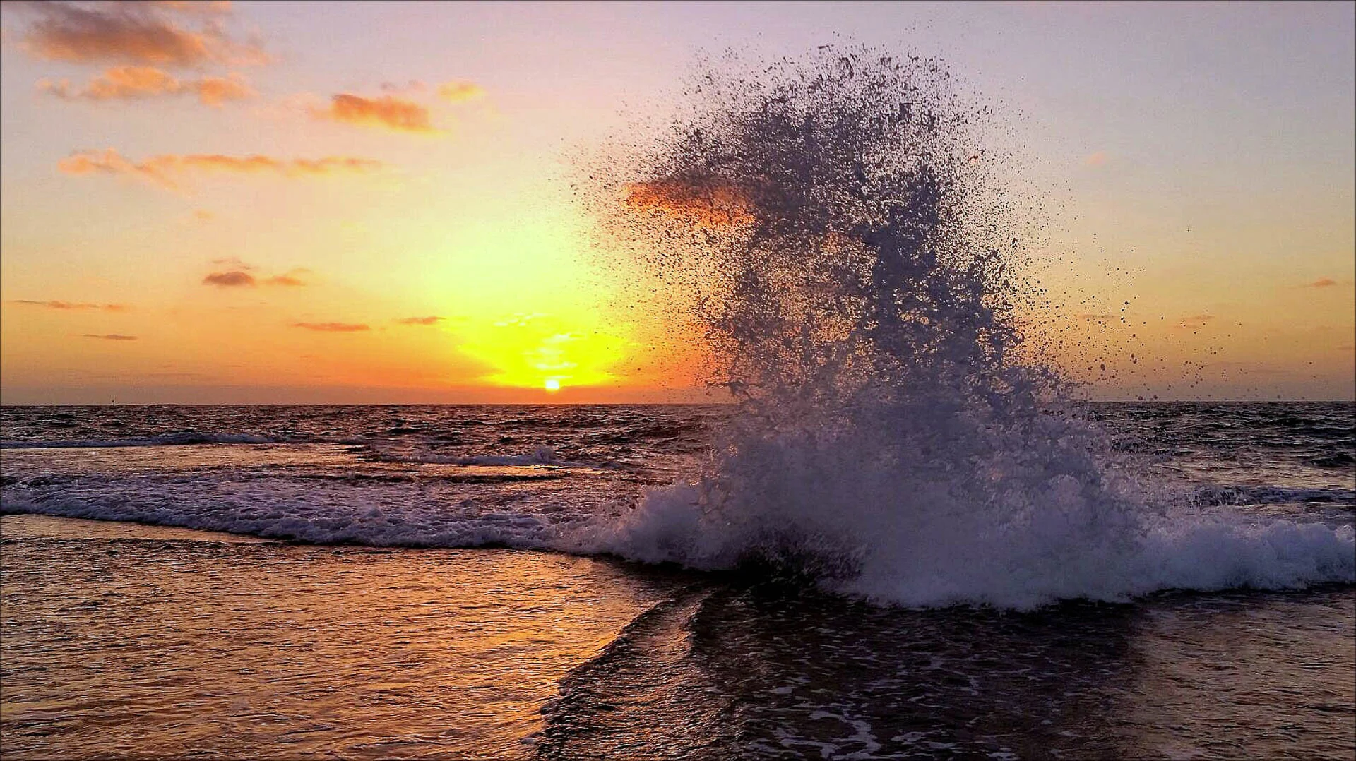 Ocean wave crashing at sunset with colorful sky and sun near the horizon.