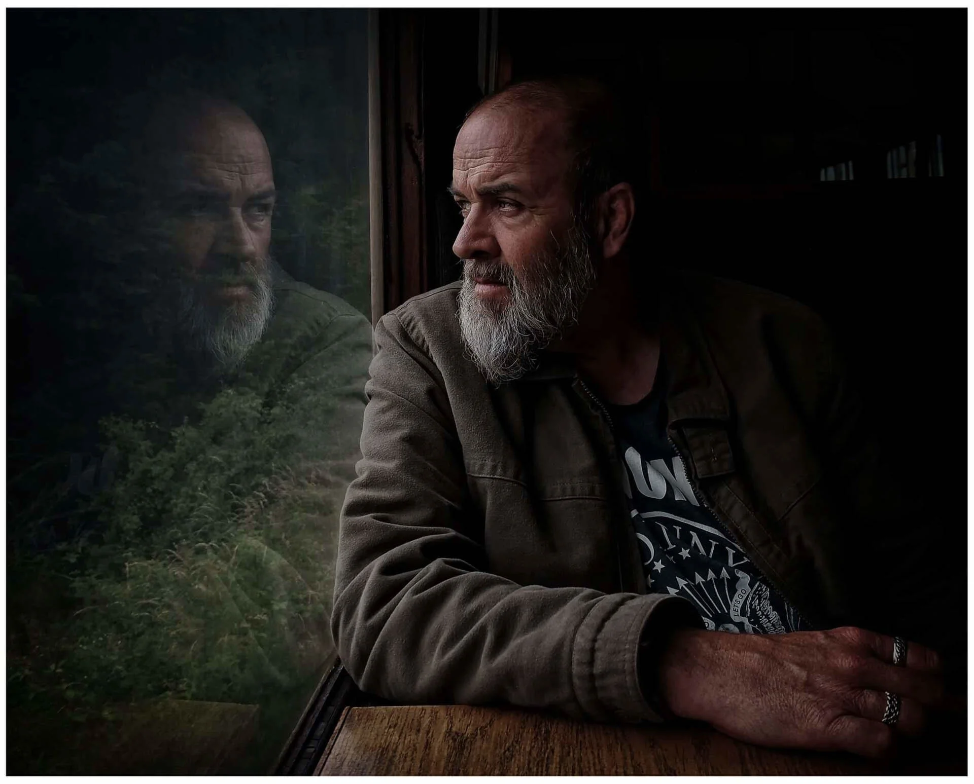 A man with a beard gazing out of a window, with his reflection visible on the glass. He is wearing a jacket and rings on his fingers. The background outside the window appears to be natural scenery.