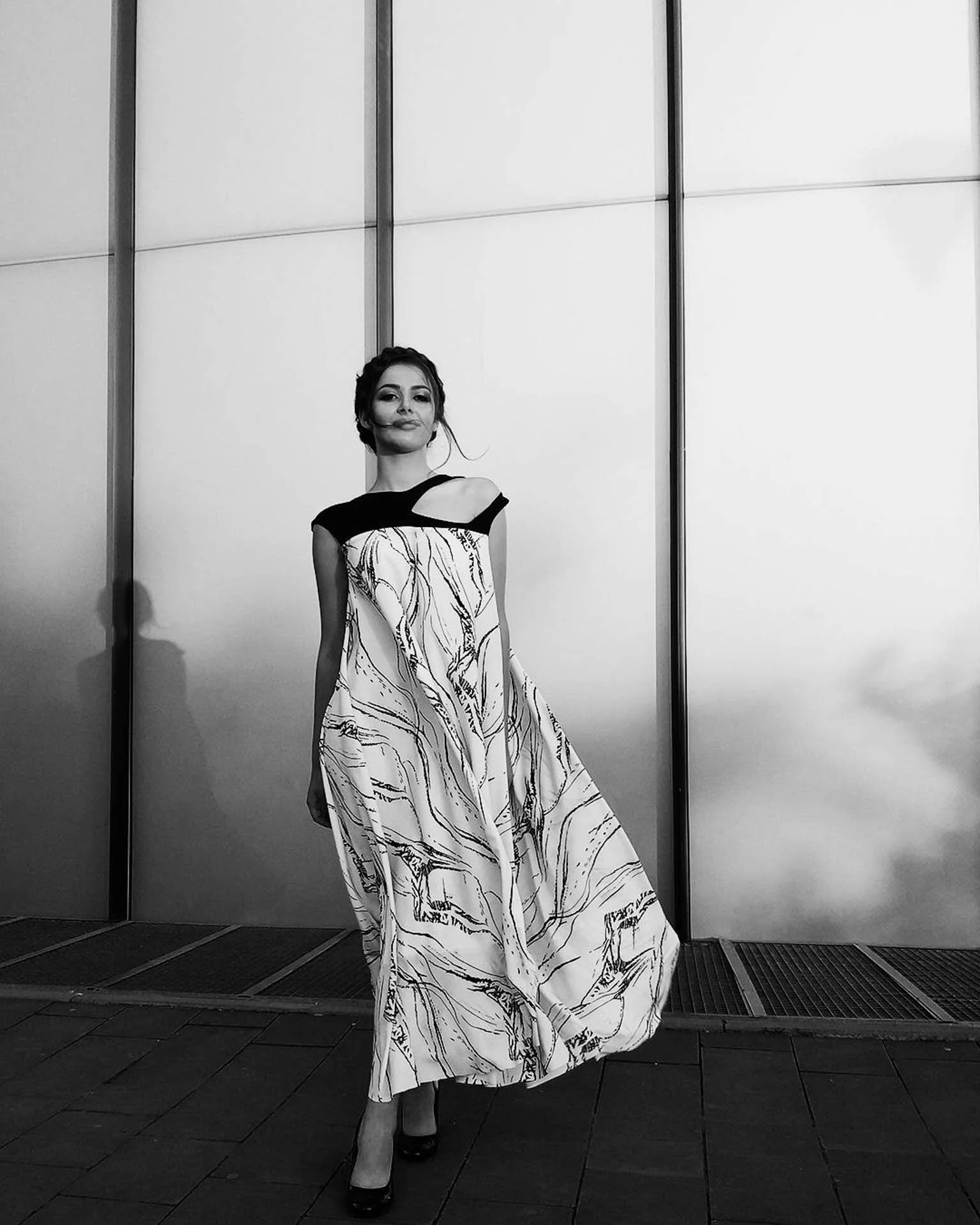 Woman in a black and white patterned dress walking on a paved path in front of a minimalist wall. Black and white photo.