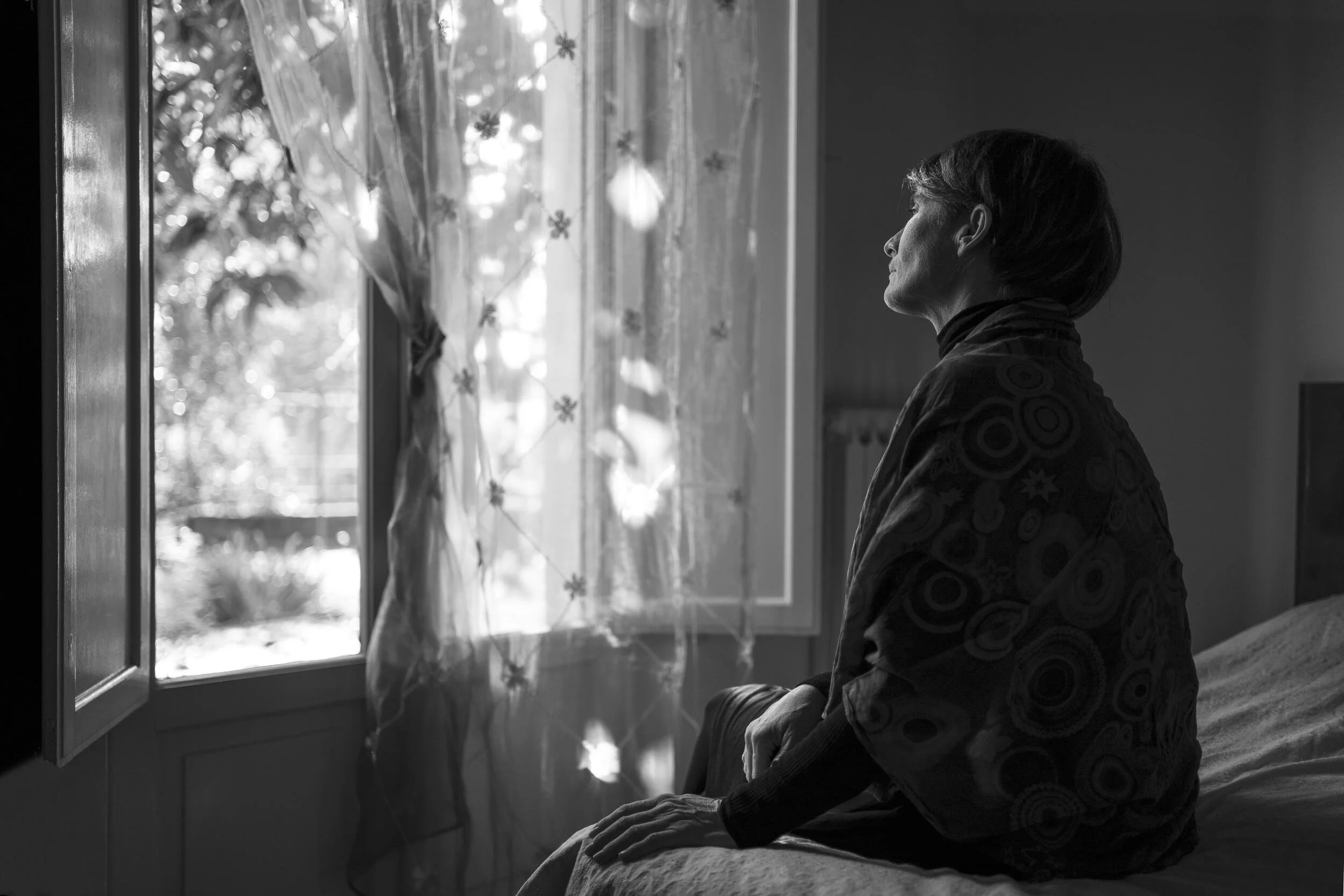 Black and white photo of a person wrapped in a blanket, sitting on a bed, gazing out a sunlit window with sheer curtains.