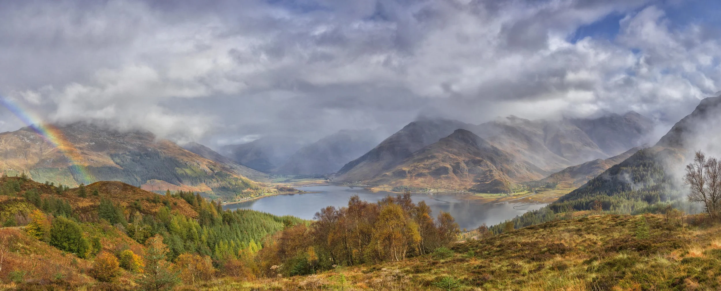 Scenic view of a lush valley with a lake, surrounded by mountains, under cloudy skies. A rainbow is visible over the mountainous terrain, adding color to the landscape.