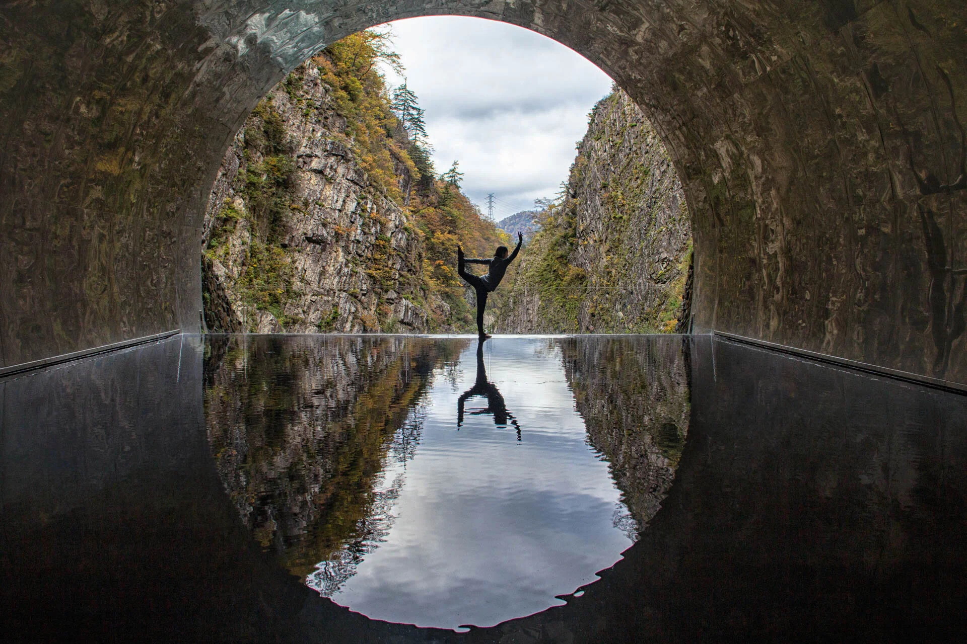 Silhouette of a person performing a yoga pose on a reflective water surface inside a tunnel, with a view of rocky cliffs and a cloudy sky.