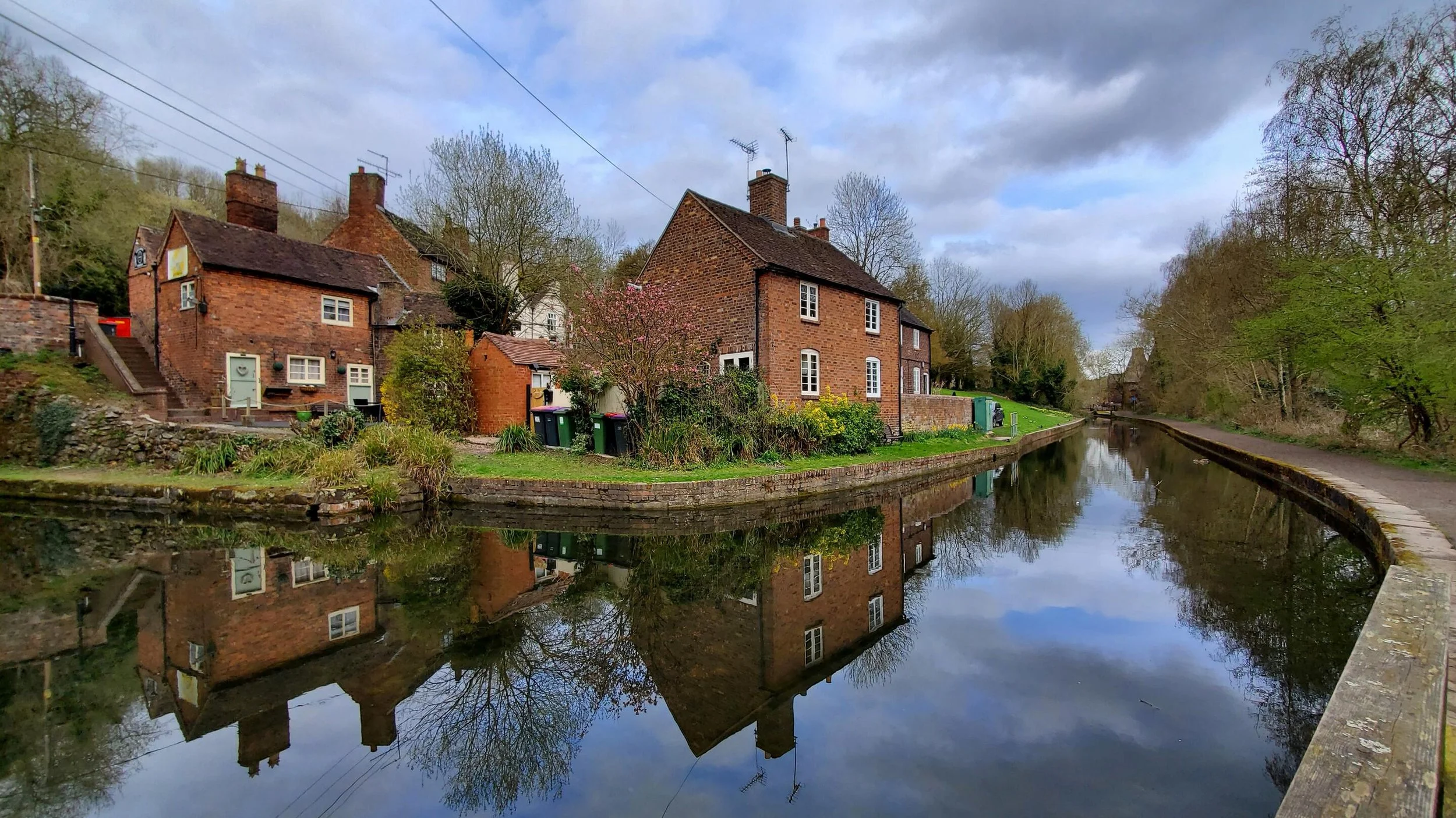 Brick houses along a canal, surrounded by trees and greenery, with reflections in the water under a cloudy sky.