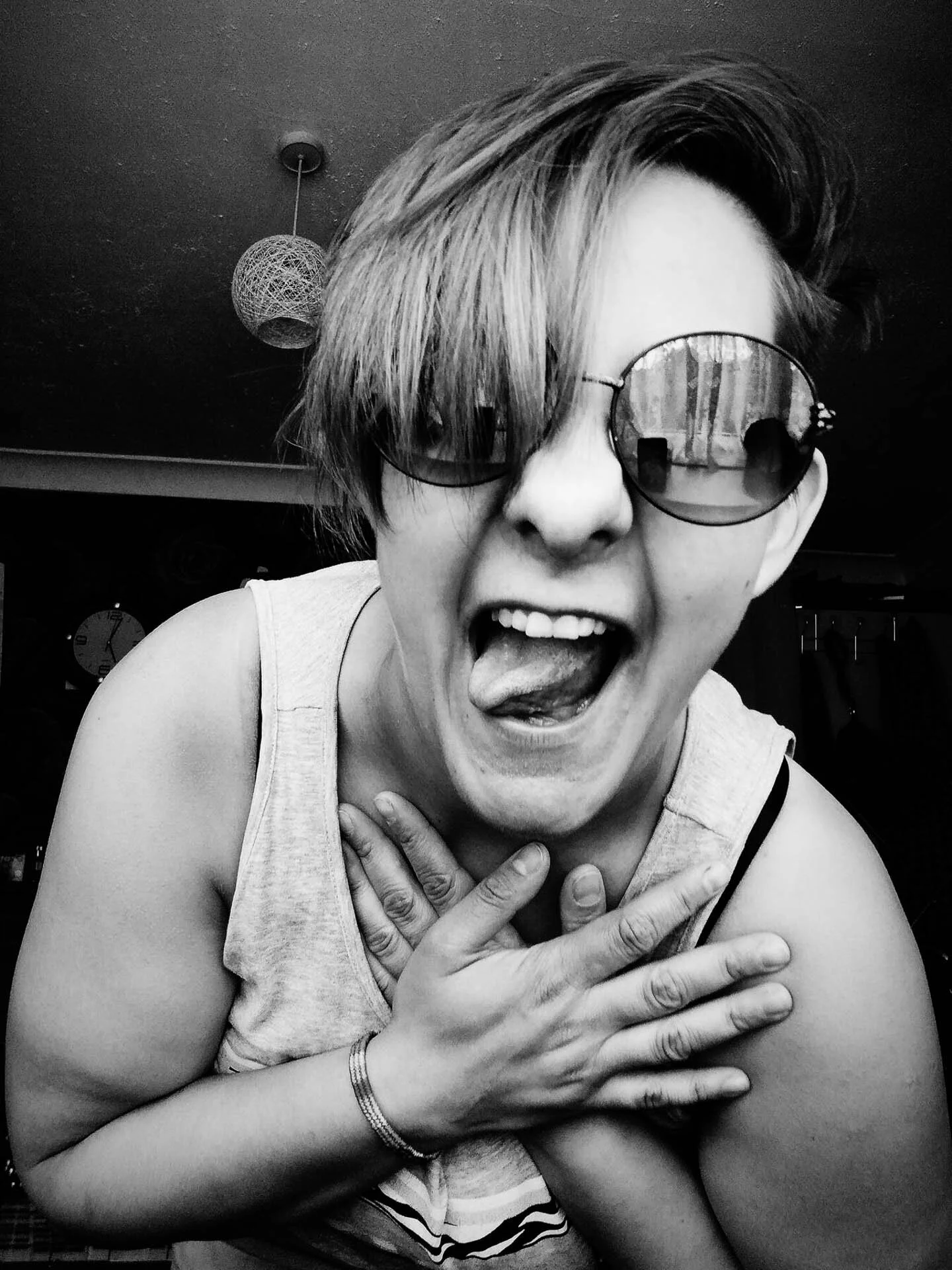 Person with short hair and sunglasses, making a playful face and covering their chest, in black and white.