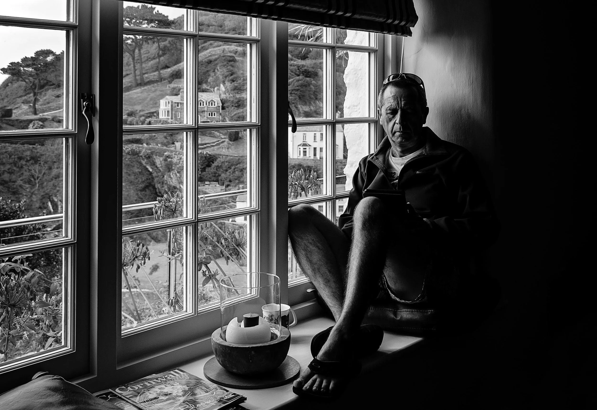 Man sitting by a window reading with a coastal view outside; items on windowsill include a candle holder, cup, and magazine.