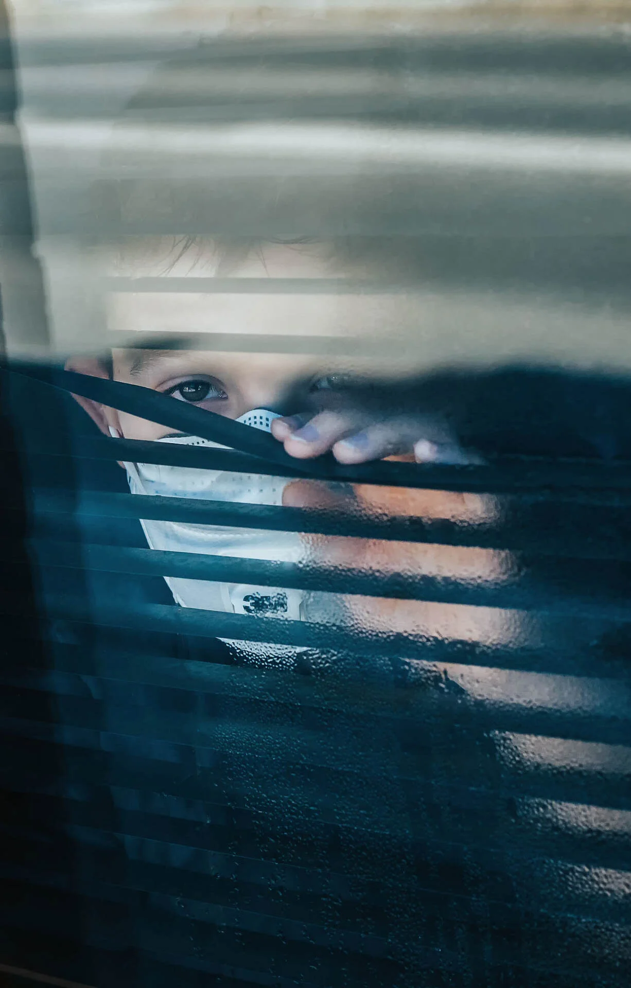 Child wearing a face mask peeking through window blinds.