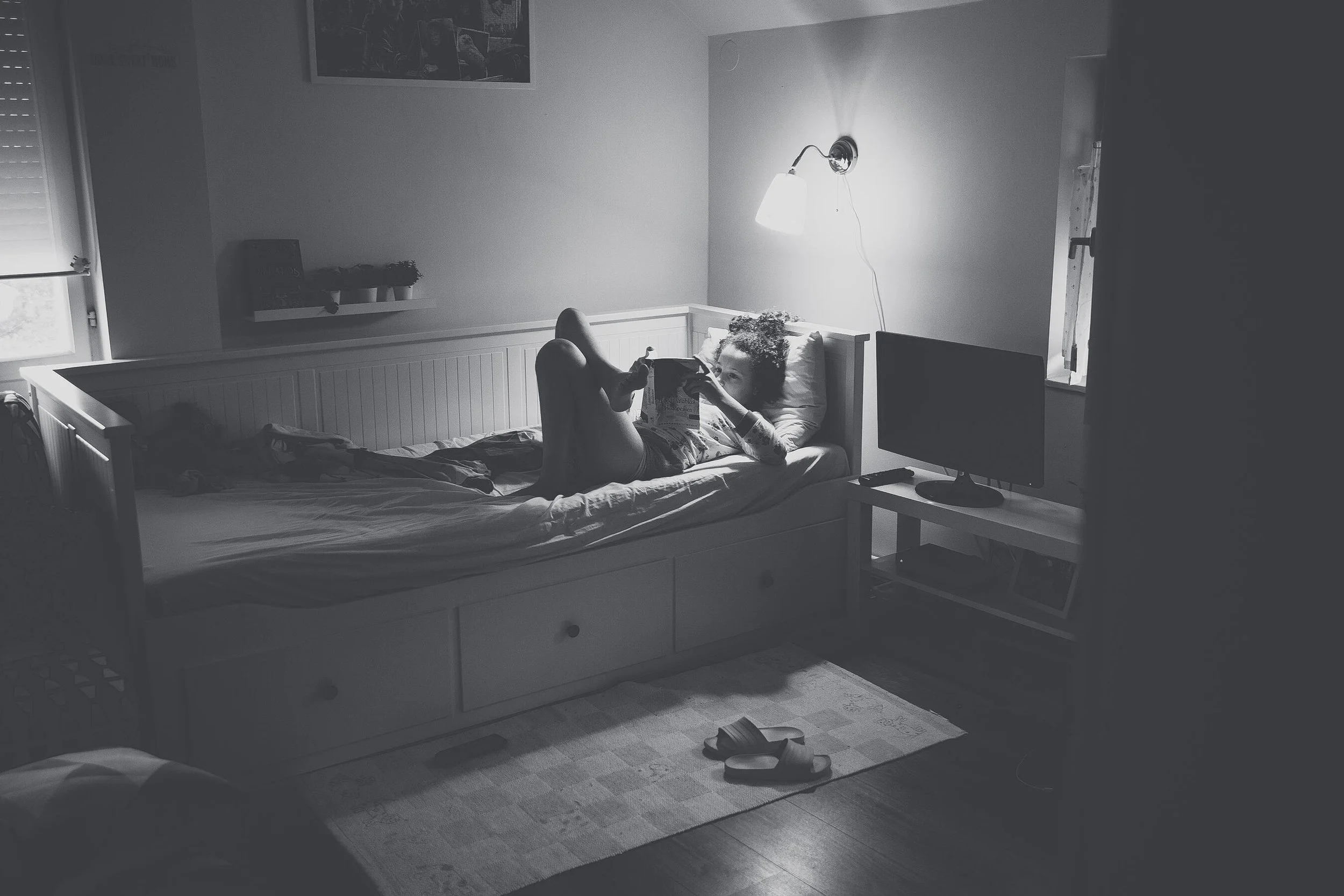 Person lying on bed reading a book in a dimly lit room with a wall lamp, TV, and slippers on the floor.