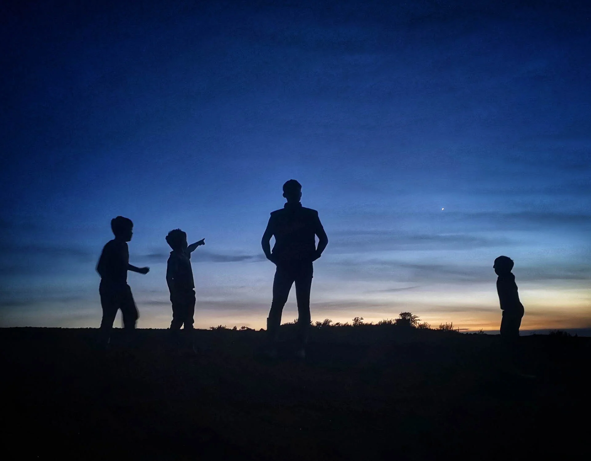 Silhouettes of four people at sunset, with one person pointing at the sky, against a twilight backdrop.
