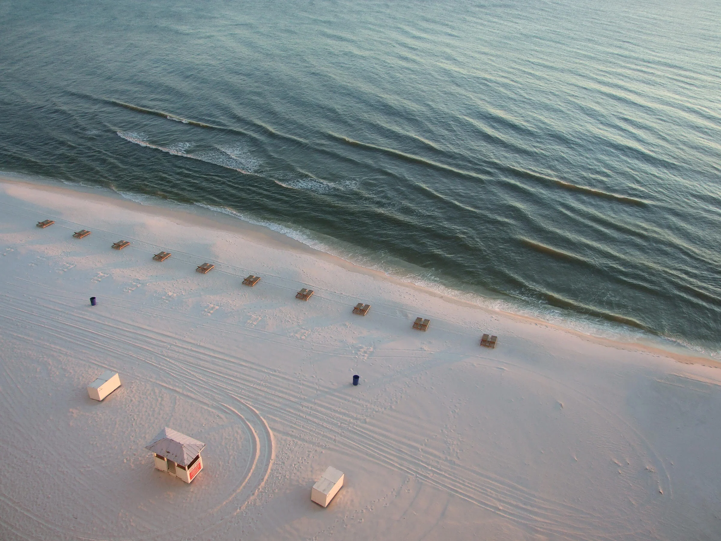 Aerial view of a sandy beach with lounge chairs arranged in a row near the ocean, and small huts on the sand. Wavy greenish-blue water approaches the shore.