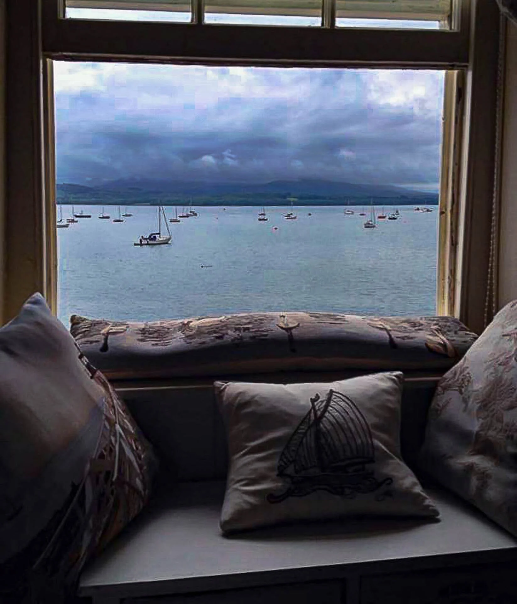 View from a window seat with cushions, overlooking a bay with sailboats and cloudy sky.