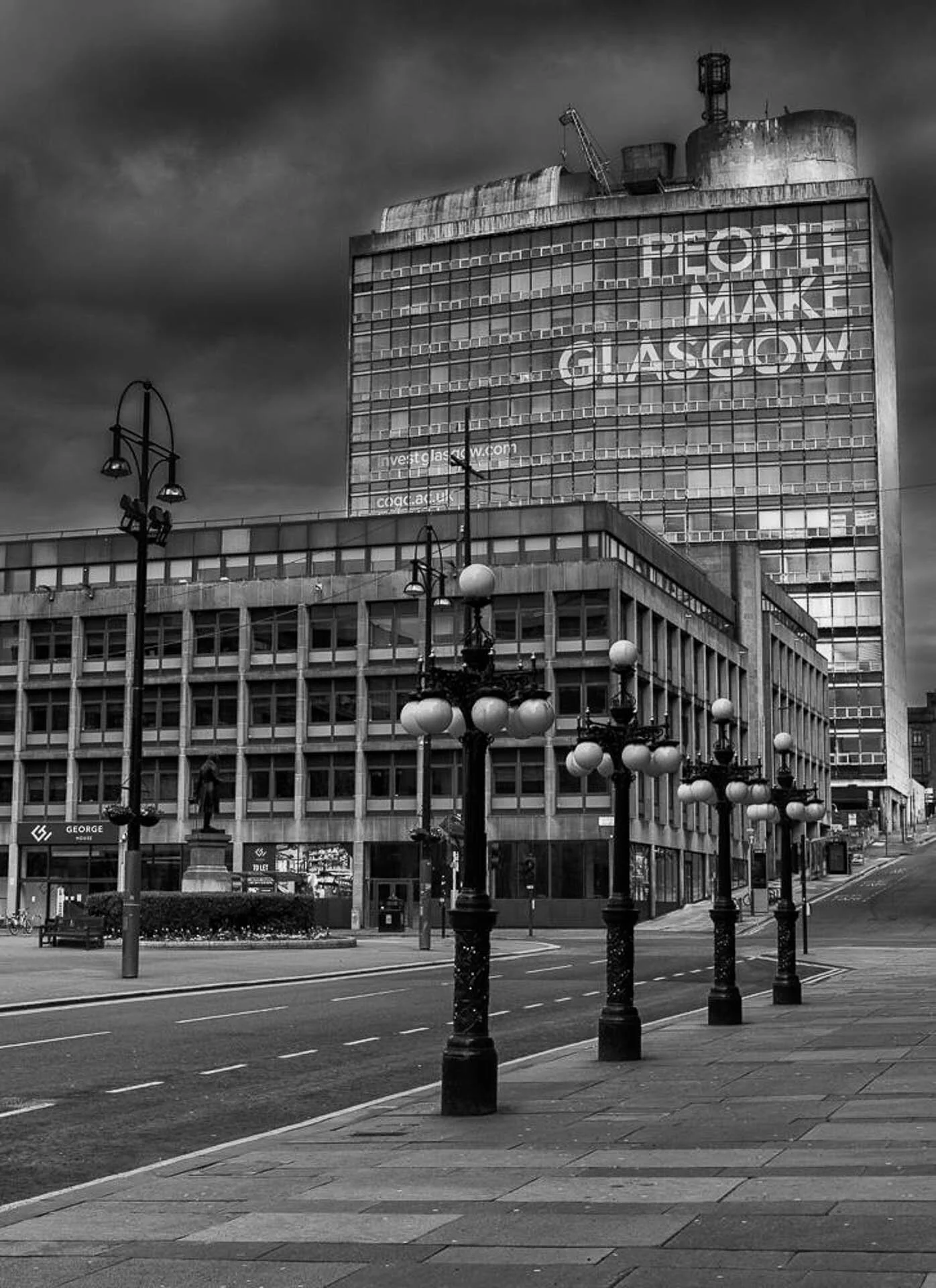 Black and white image of a tall building with "People Make Glasgow" written on it, set against a cloudy sky. The foreground features ornate street lamps and a deserted street.
