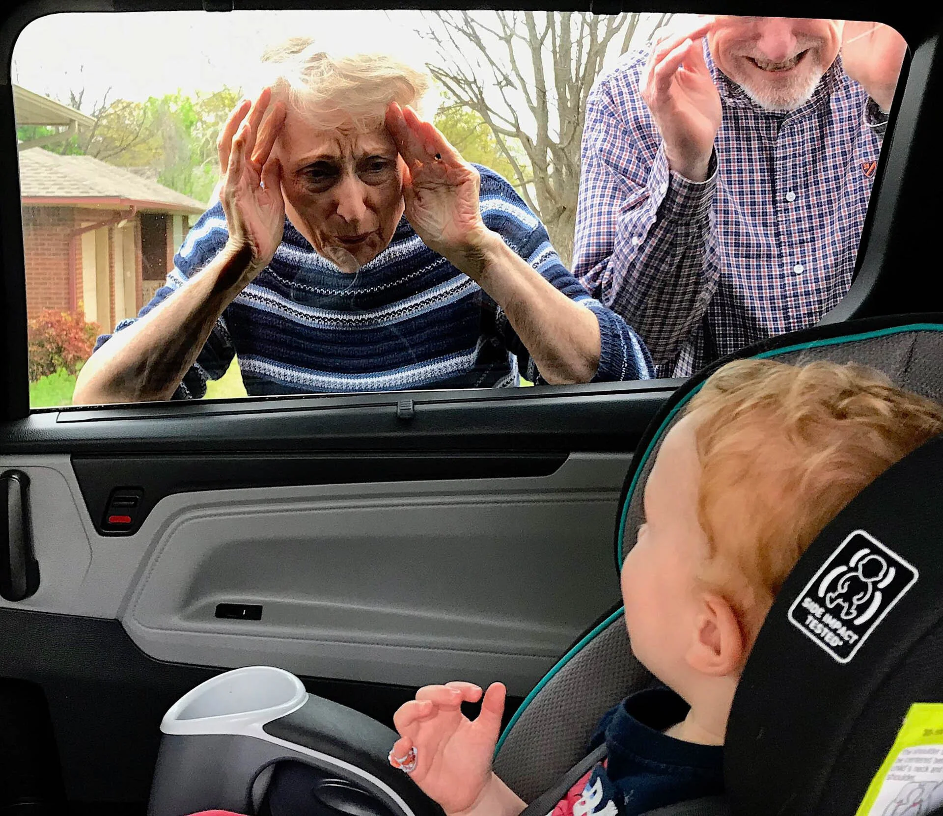 Two elderly people looking through a car window at a young child in a car seat.