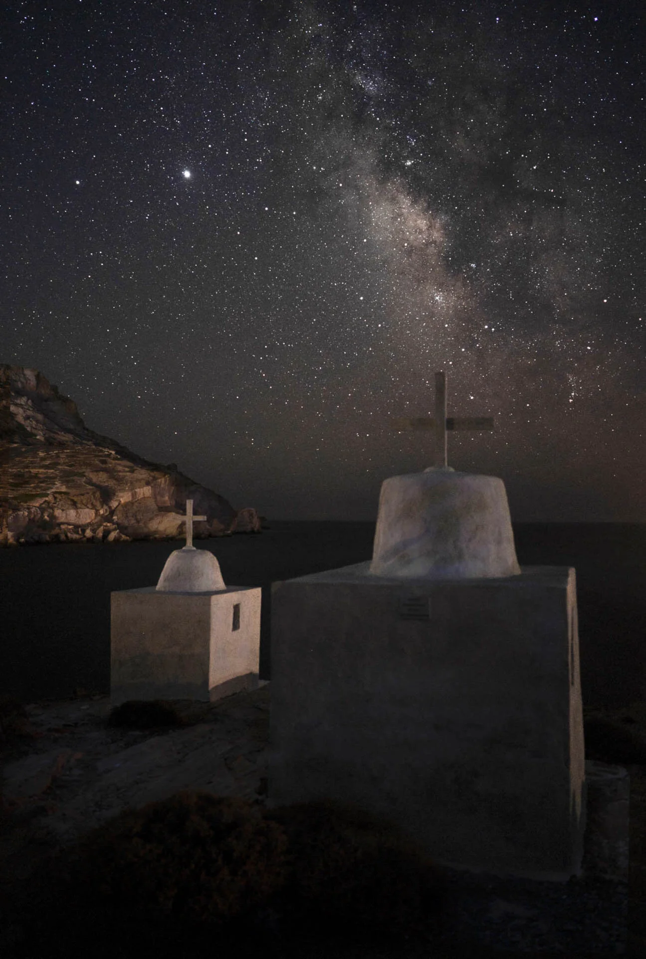 Night sky with Milky Way over two small chapels with crosses, rocky landscape in background.