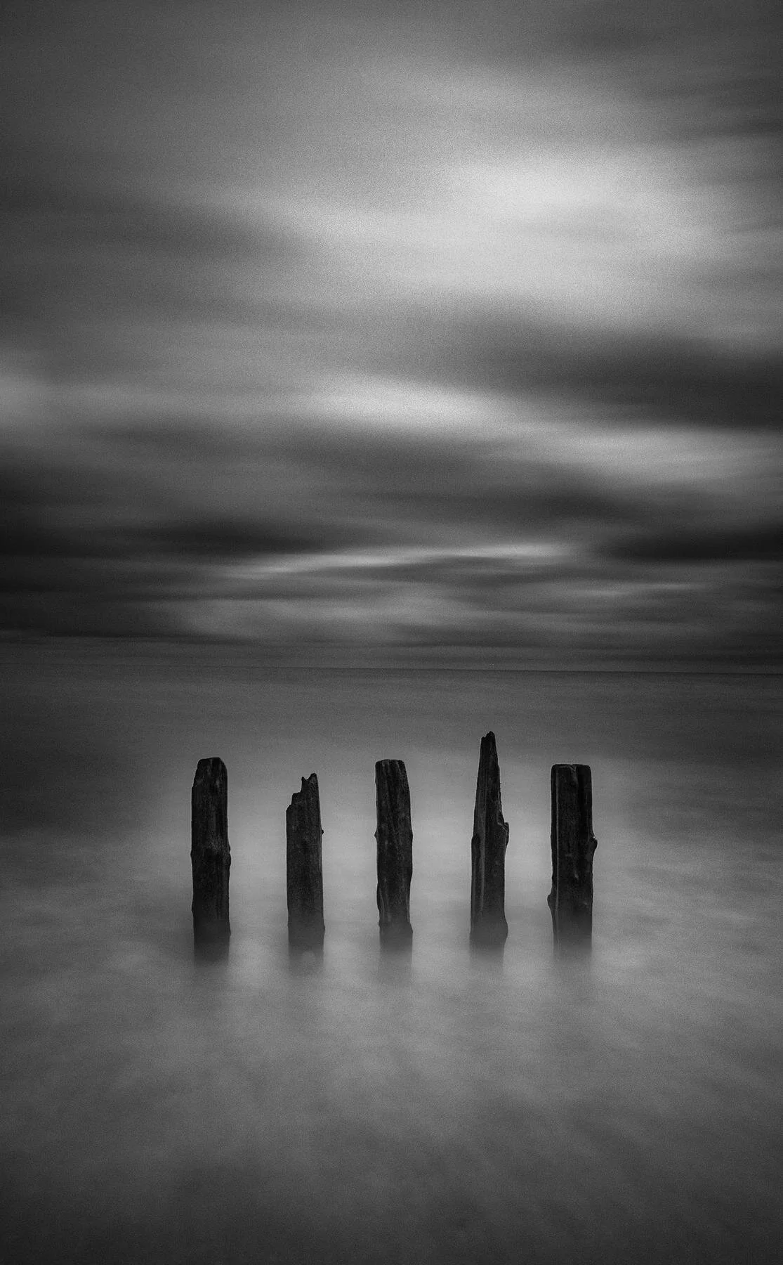 Black and white photo of five weathered wooden posts standing in calm water with a blurred cloudy sky above.