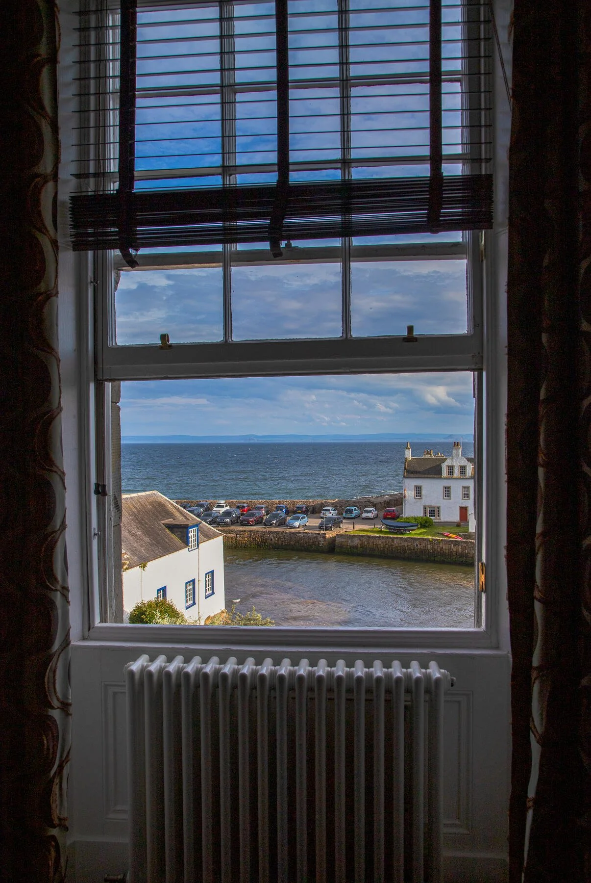 View of a seaside village through a window with blinds, featuring buildings near the water, parked cars along the shoreline, and a clear blue sky.
