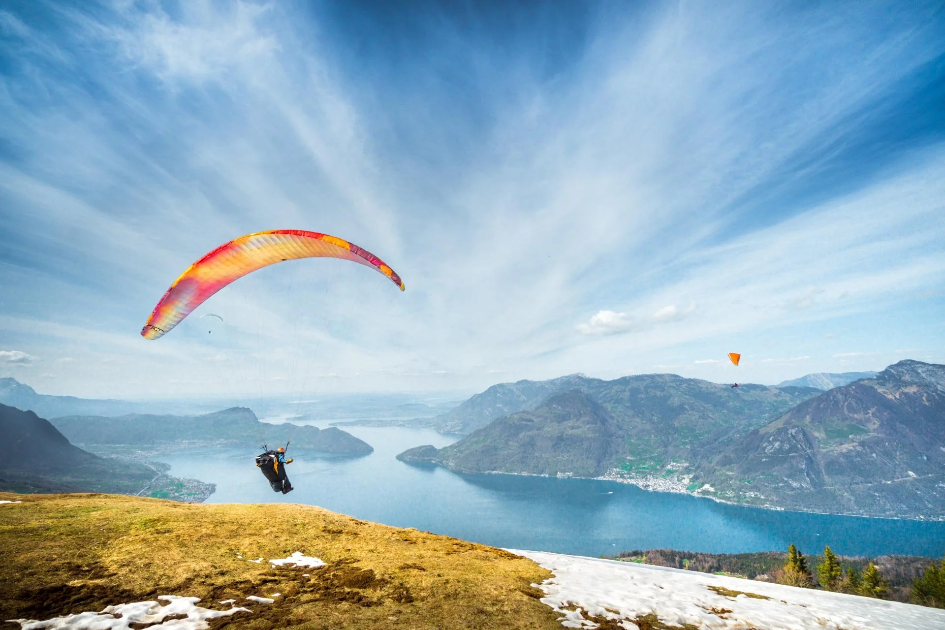 Paraglider flying over a scenic mountain landscape with a lake and snow patches on the ground.