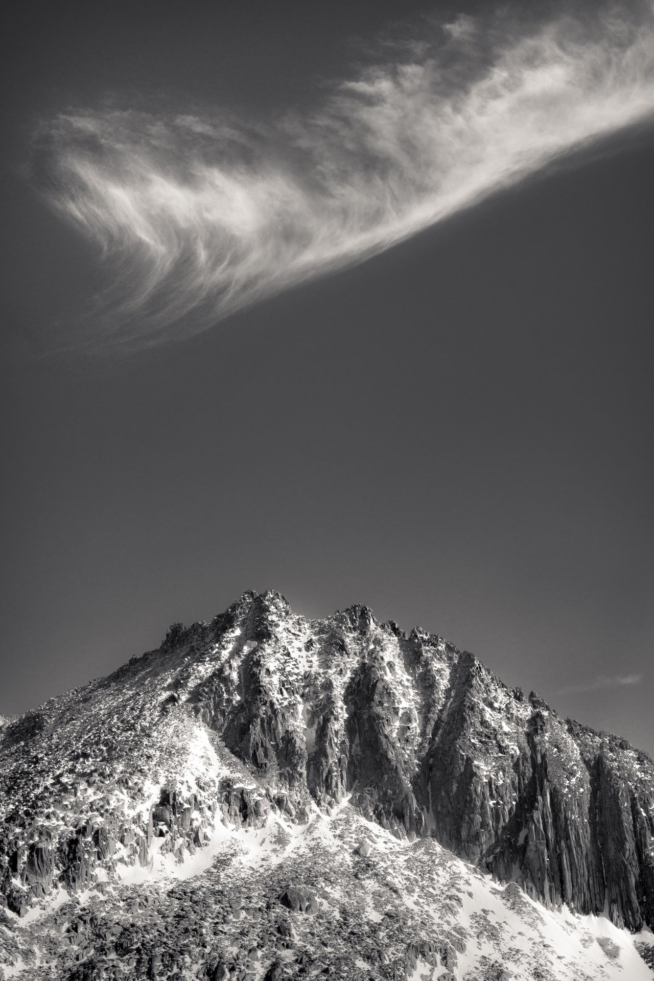 Snow-covered mountain peak with jagged rocks under a streaky cloud in a clear sky.