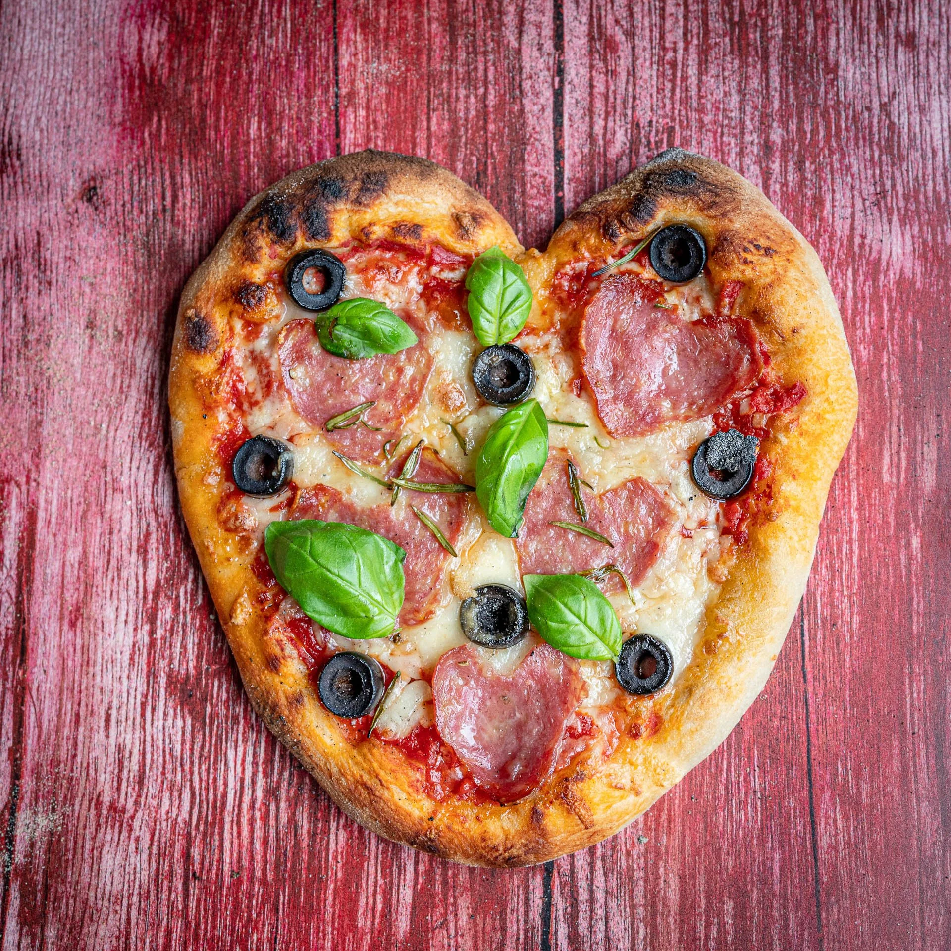 Heart-shaped pizza with salami, black olives, basil leaves, and cheese on a red wooden table.